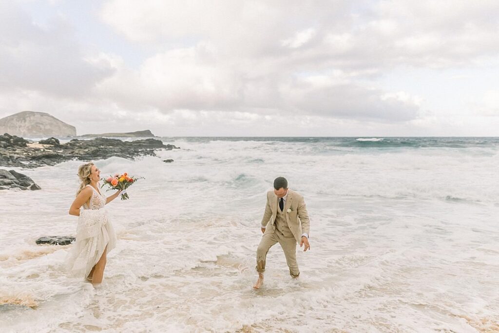 A bride and groom walk through the water on an Oahu beach 