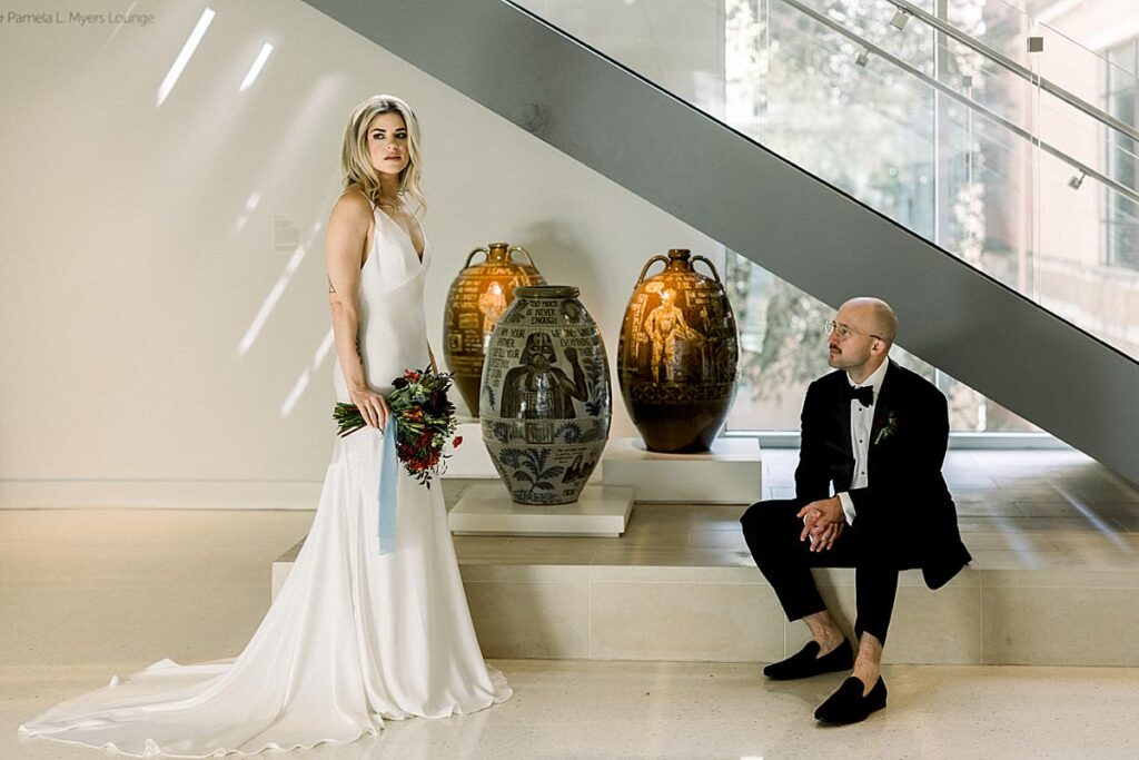 A bride stands under some stairs by some vases while her groom sits on a step at their Asheville Art Museum Wedding