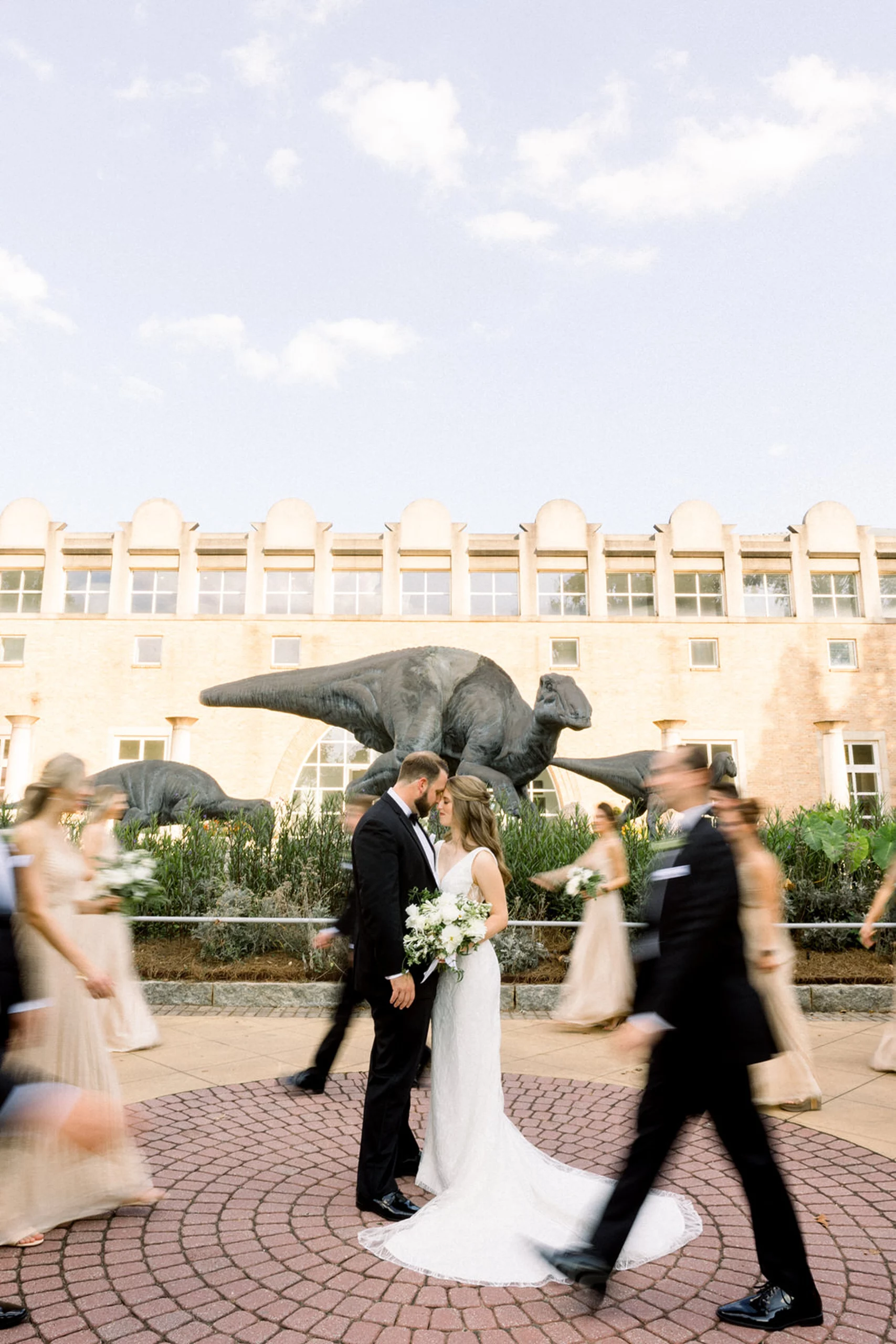 bride and groom kissing while their bridal party walks around them with a dinosaur in the background at a fernbank museum wedding