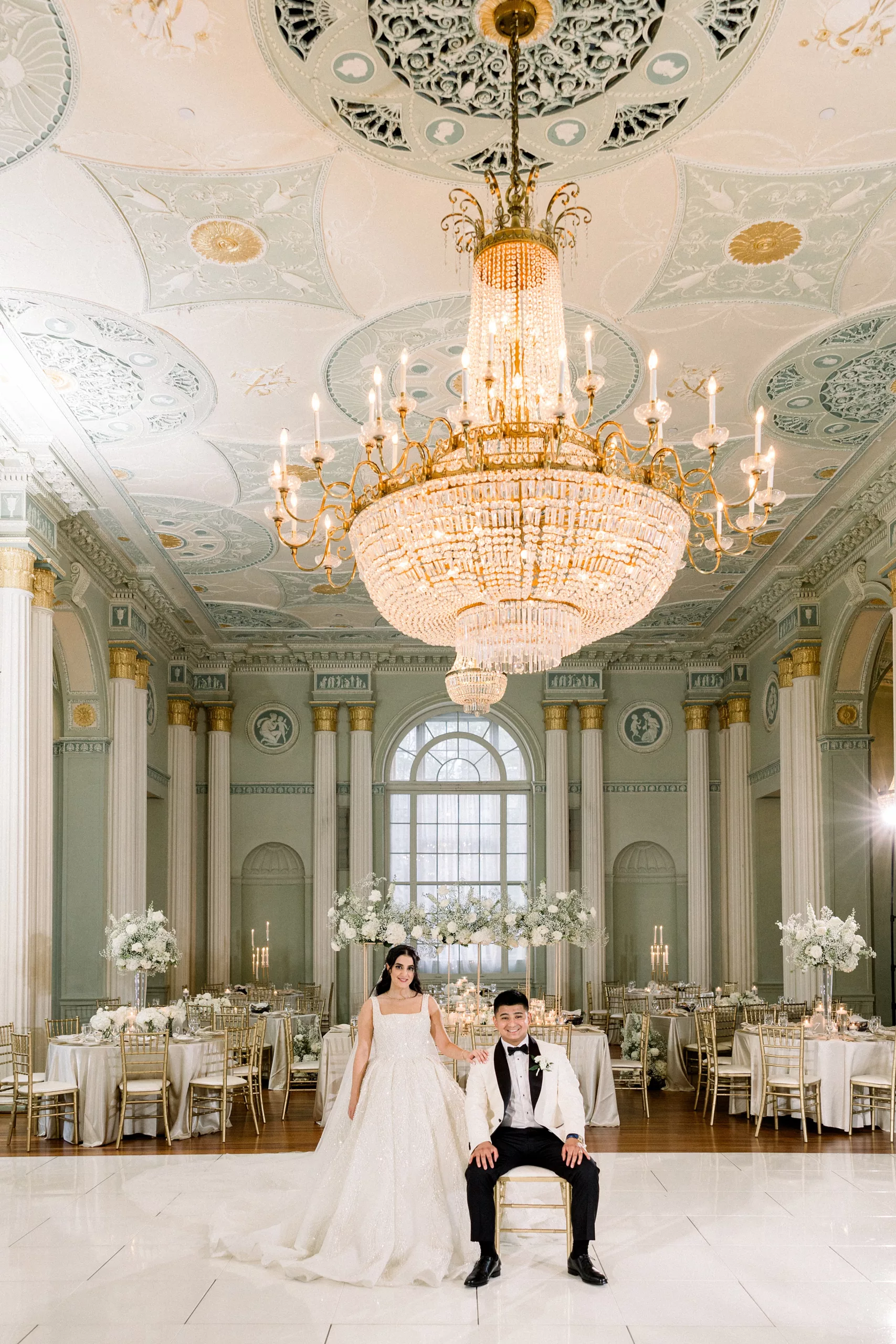 Newlyweds sit under a large crystal chandelier in their reception venue at Biltmore Ballrooms Wedding