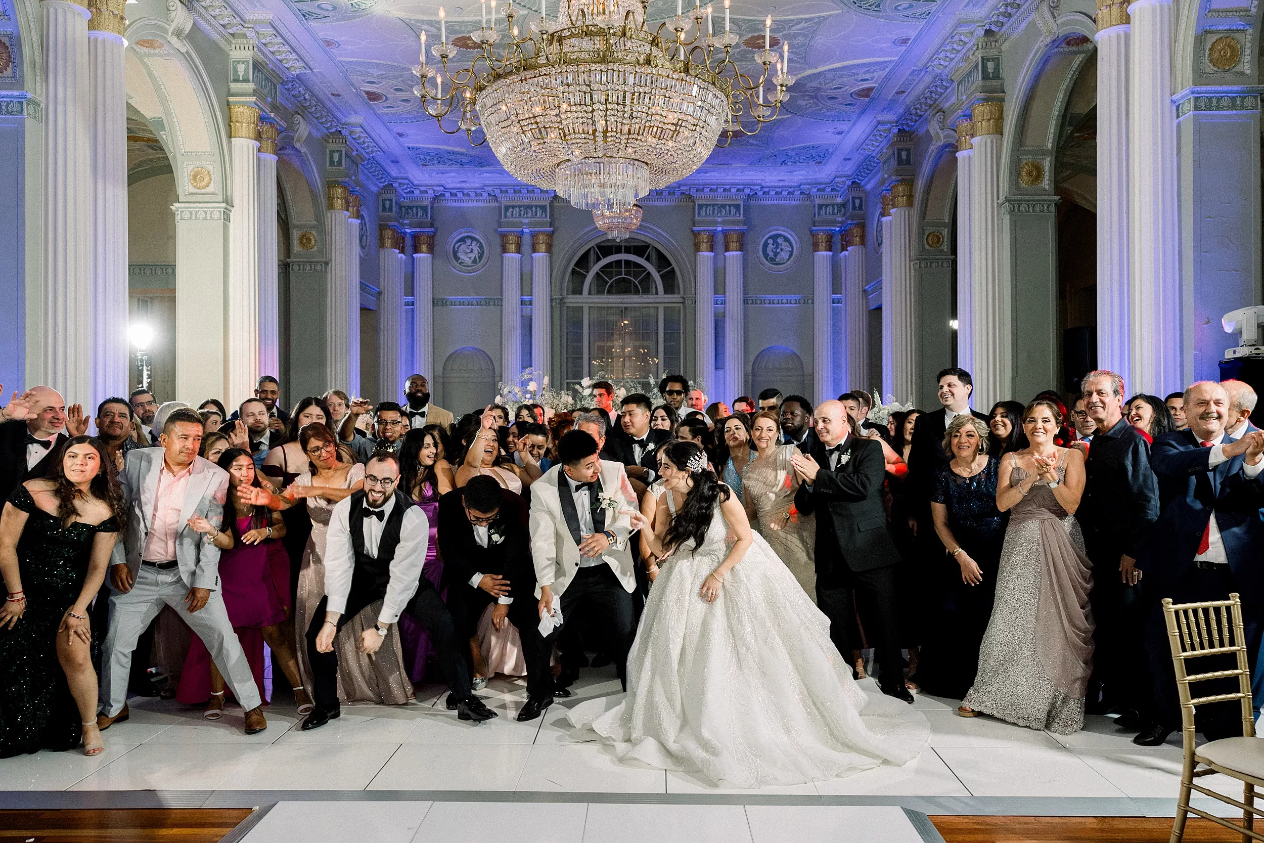 The guests of a wedding dance and celebrate behind the bride and groom under a large crystal chandelier at a Biltmore Ballrooms Wedding