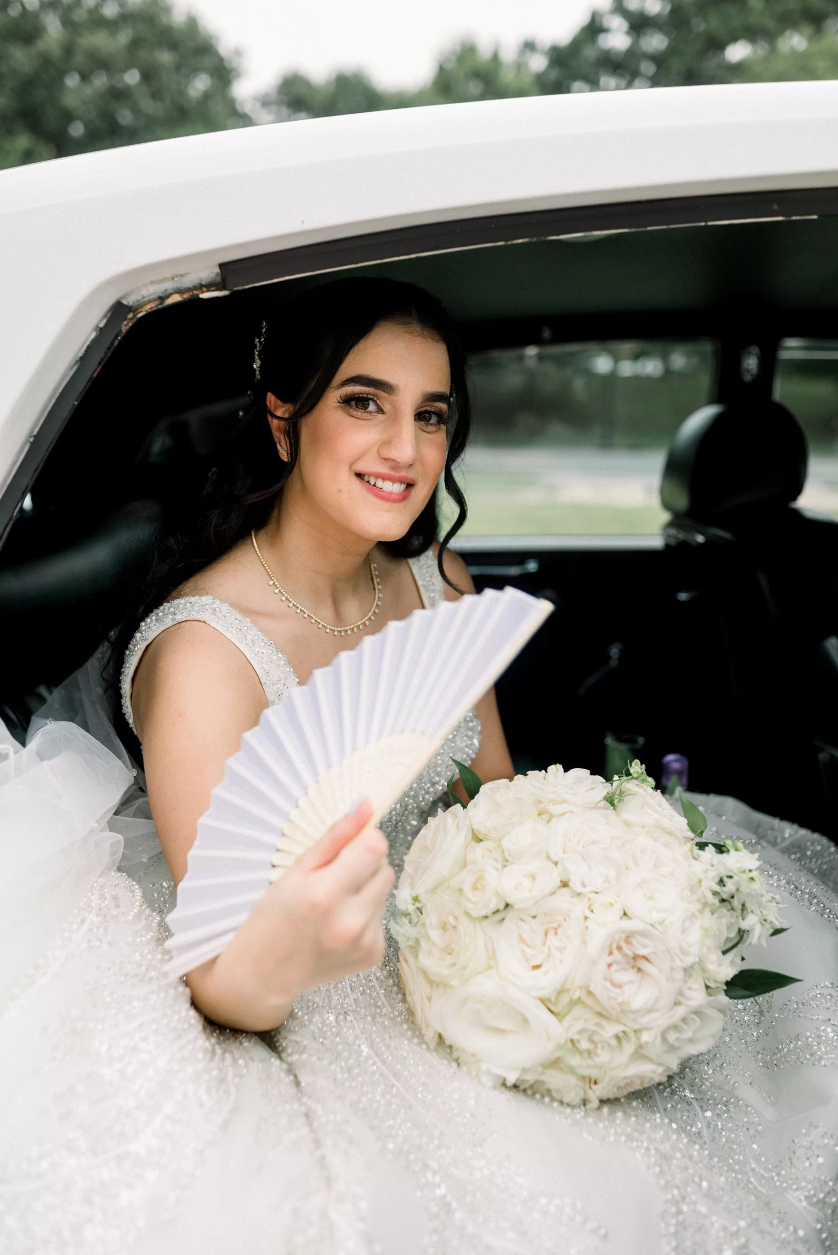 A bride fans herself while sitting in the back of a limo holding her large white bouquet