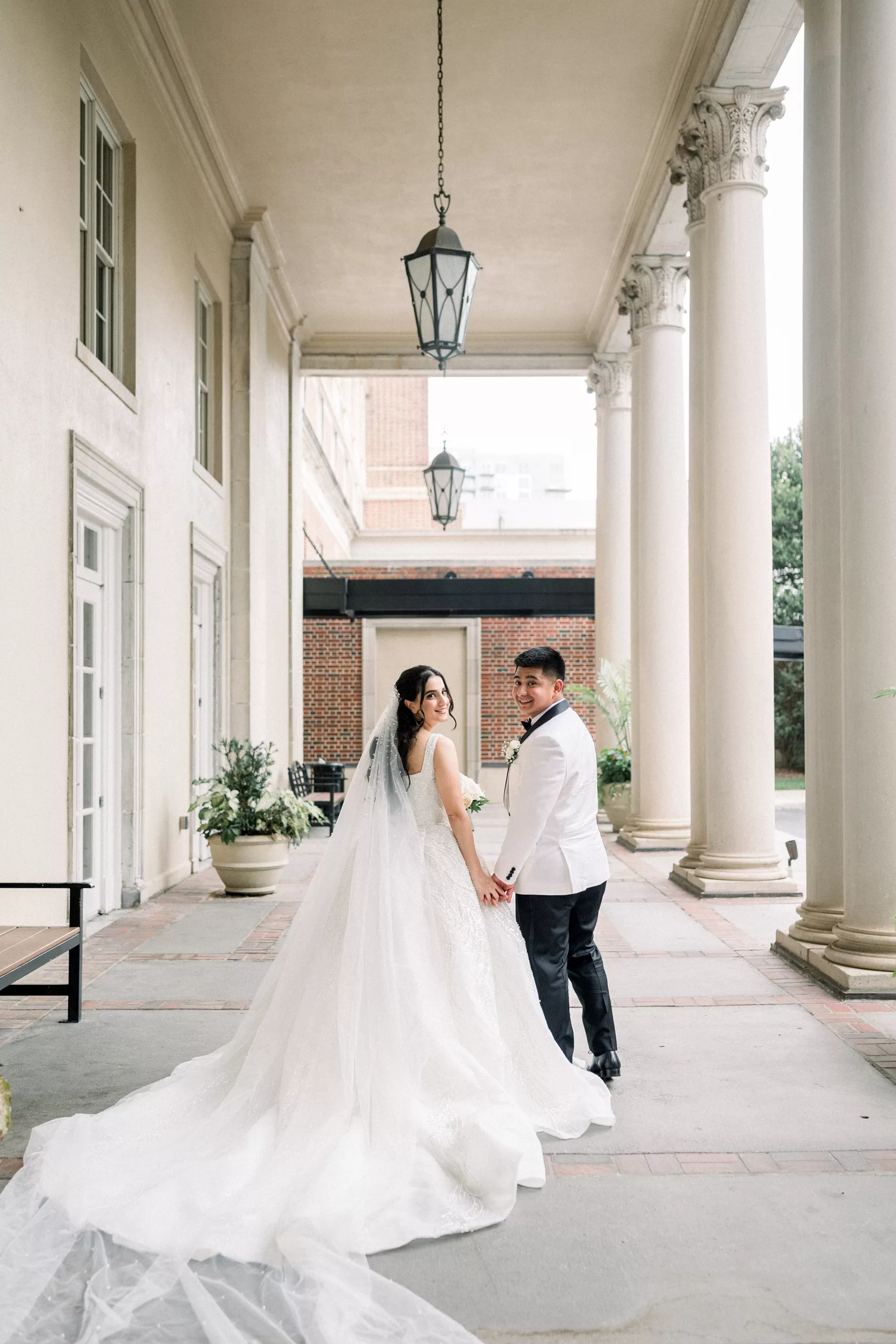 Newlyweds look over their shoulder while holding hands walking on the columned porch of the Biltmore Ballrooms Wedding venue