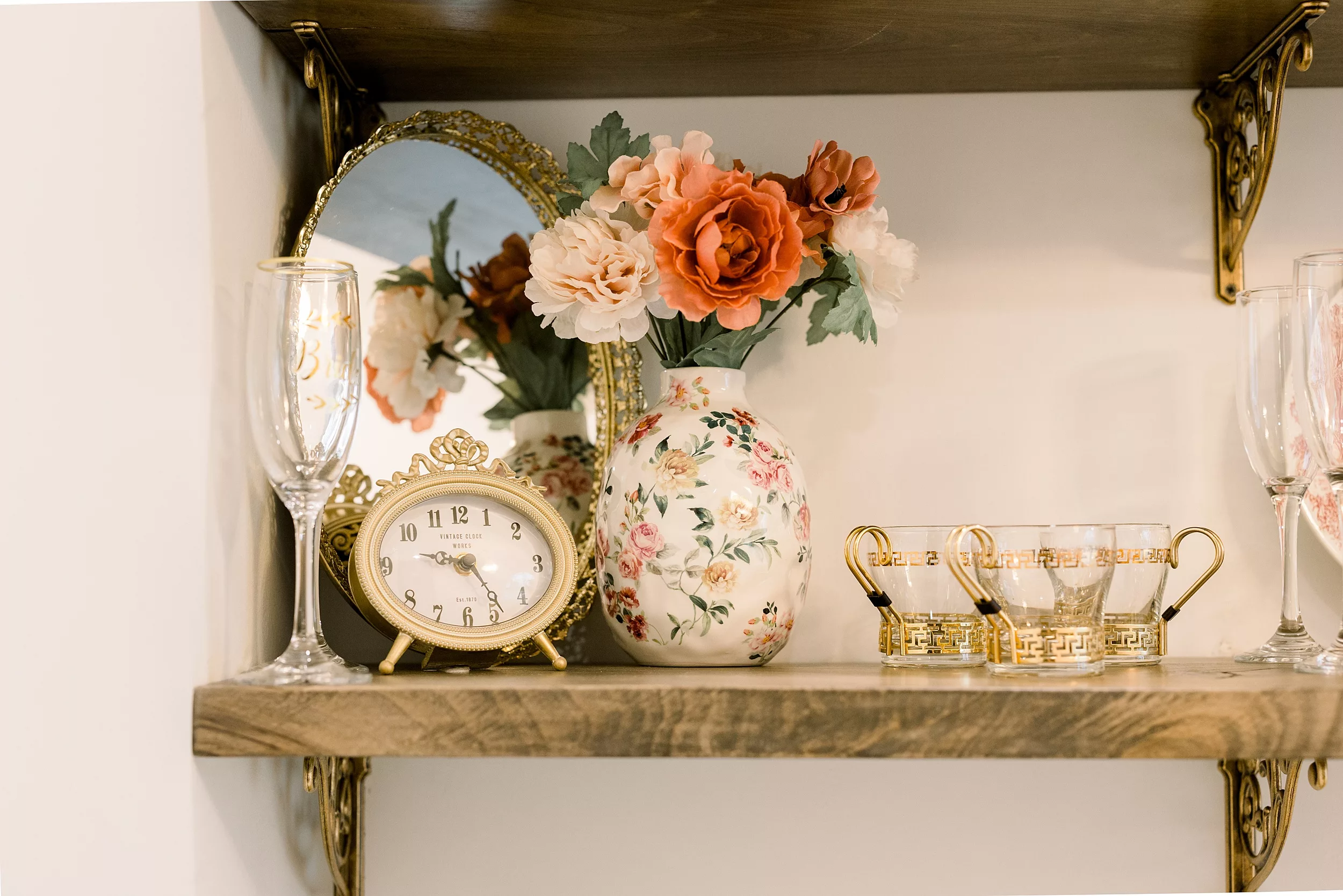 Details of an antique clock, mirror and glasses on a wooden shelf with flowers in a vase