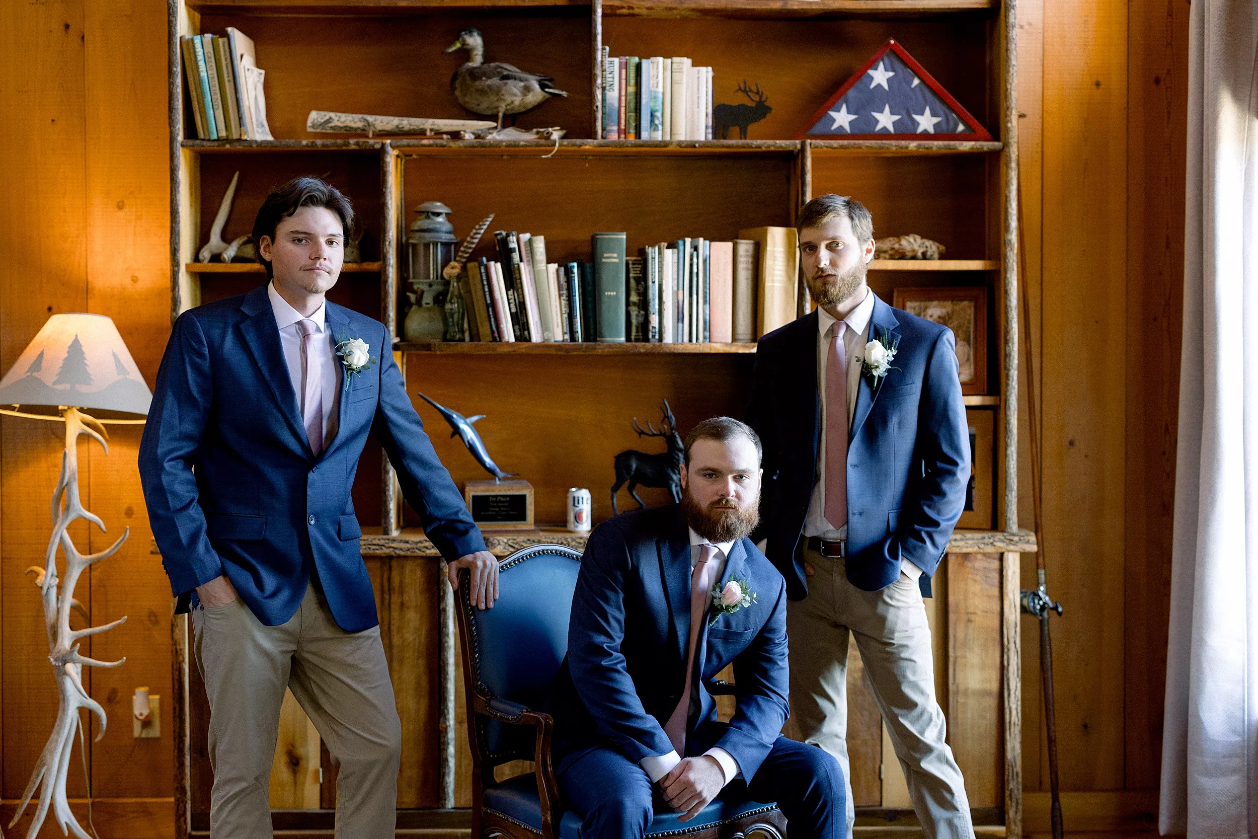 Tw men stand in a wood paneled room in blue suit jackets and khaki pants while a third sits between them in blue pants on an antique chair Blue Mountain Vineyards Wedding