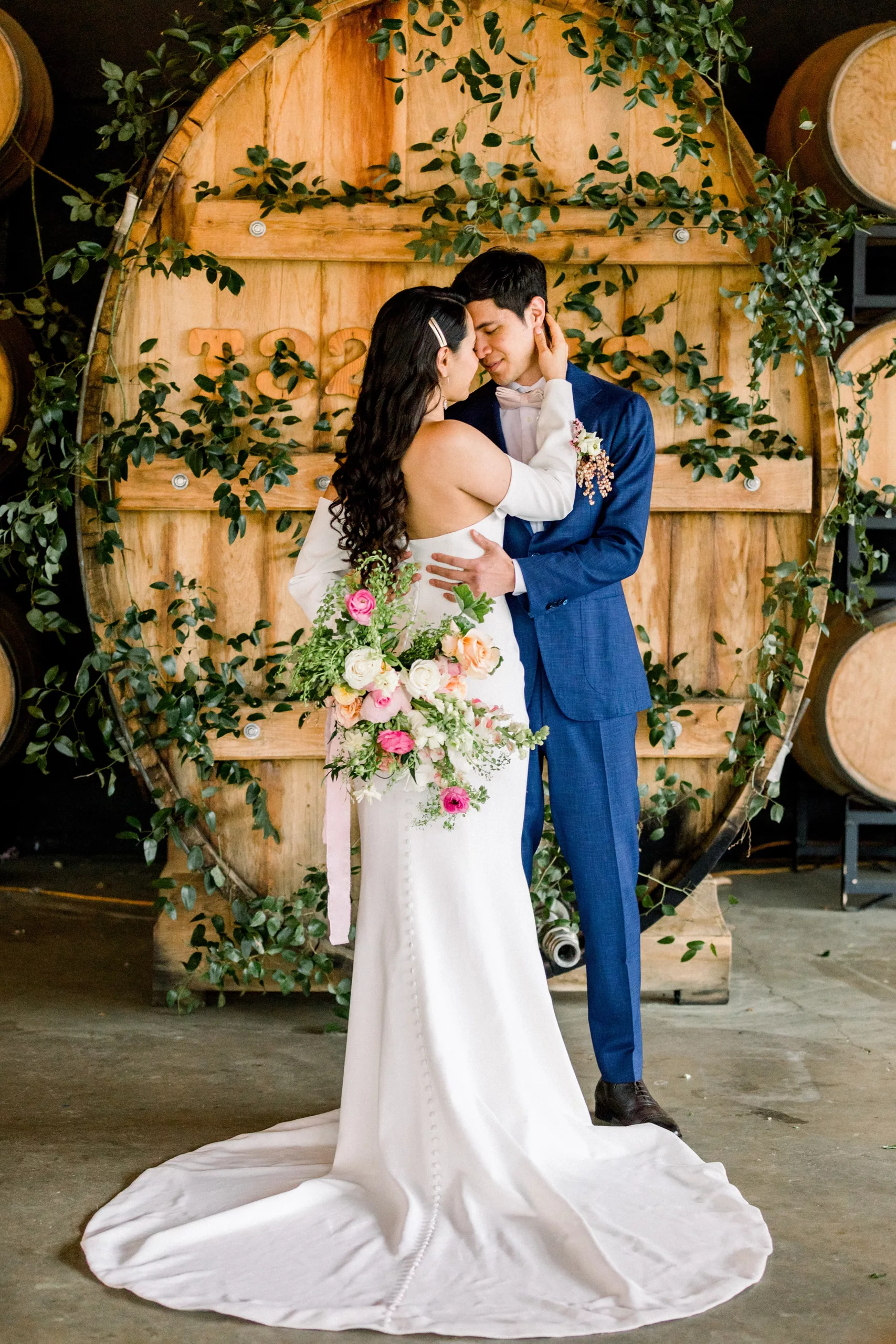 Newlyweds dance in front of a large vine covered wine barrel in a blue suit and silk white dress at a Chateau Elan Wedding