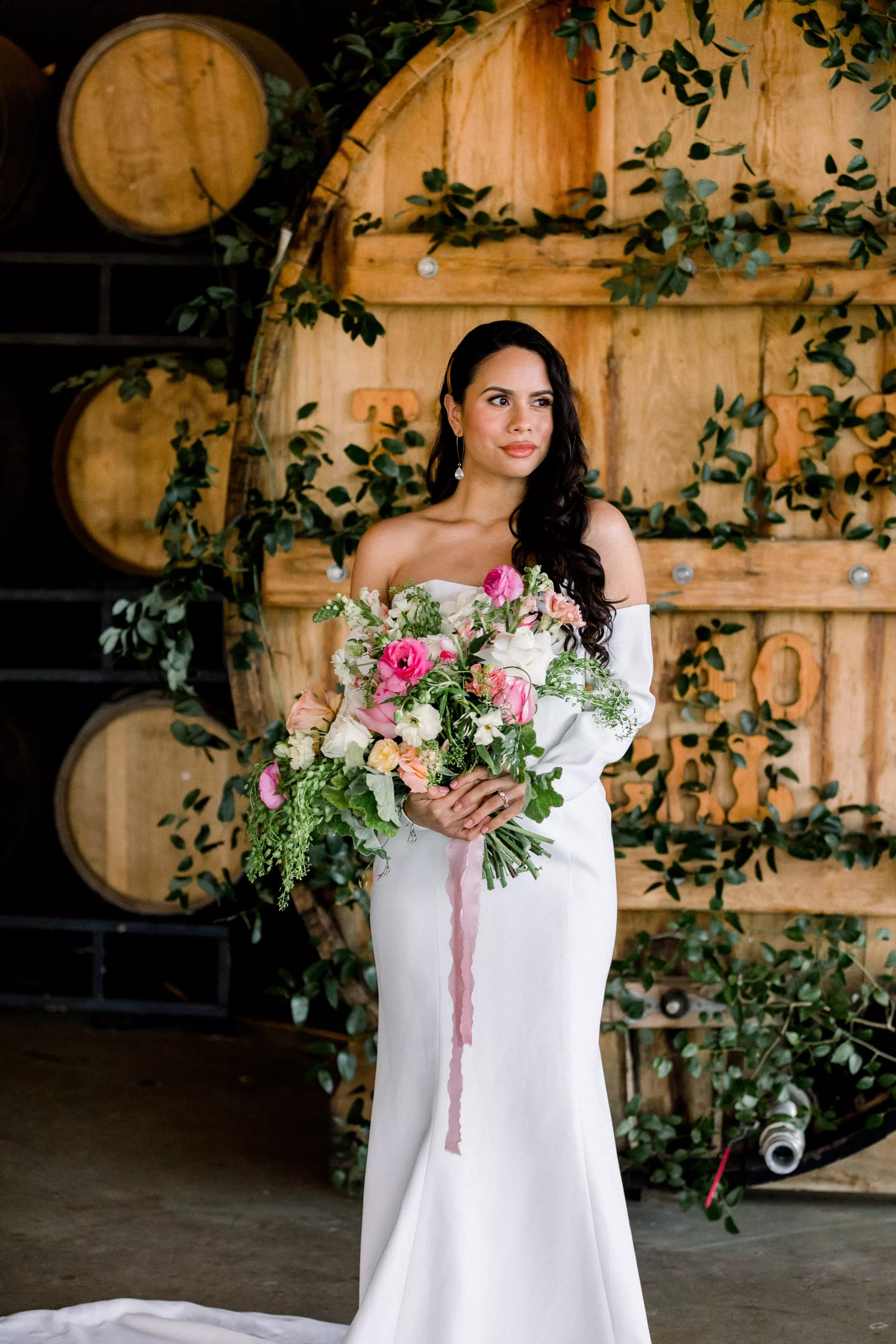 A bride in a silk dress stands in front of a large wine cask holding a large colorful bouquet at a Chateau Elan Wedding