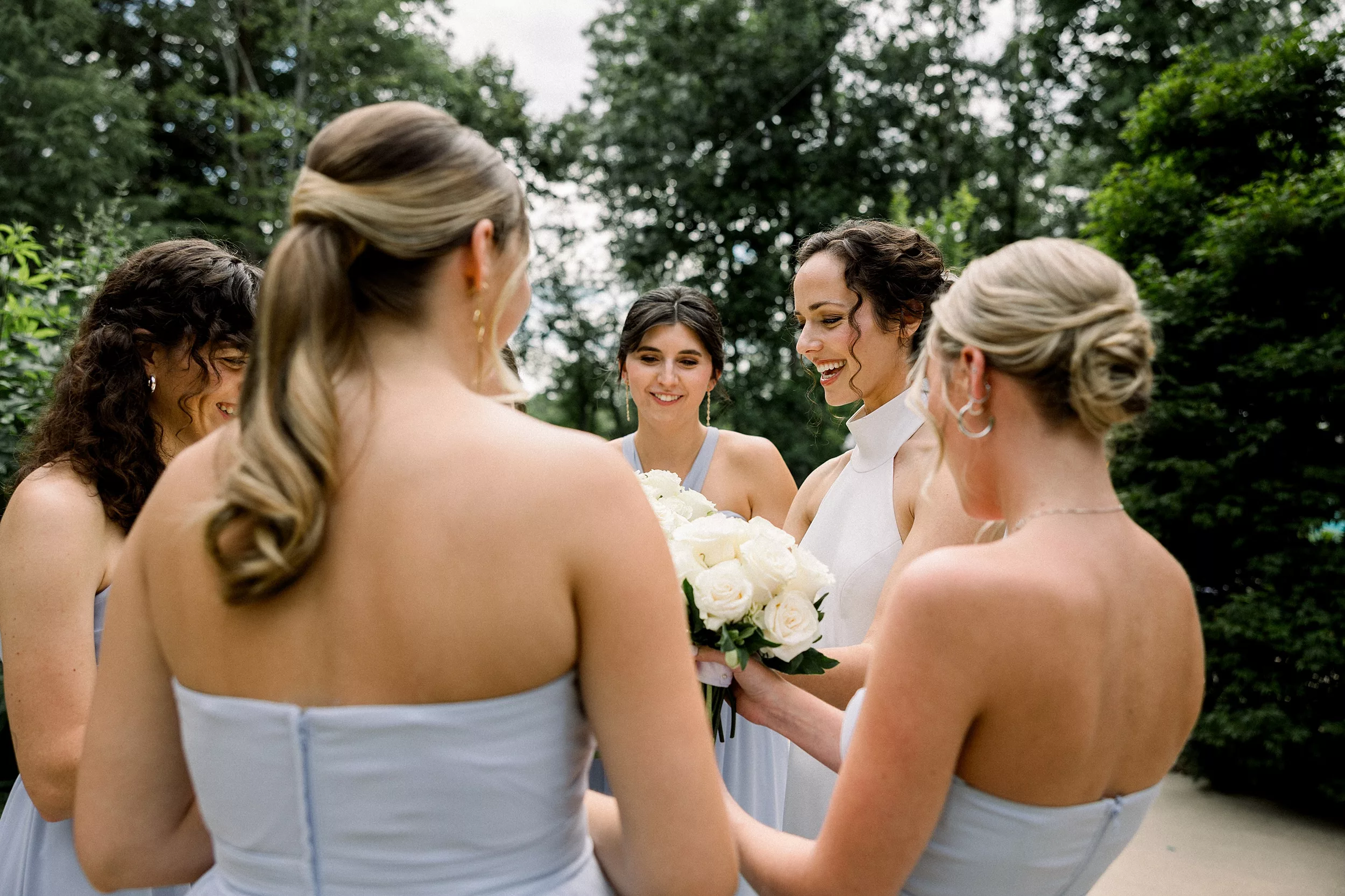 A bride and her bridesmaids smile down her her large white rose bouquet