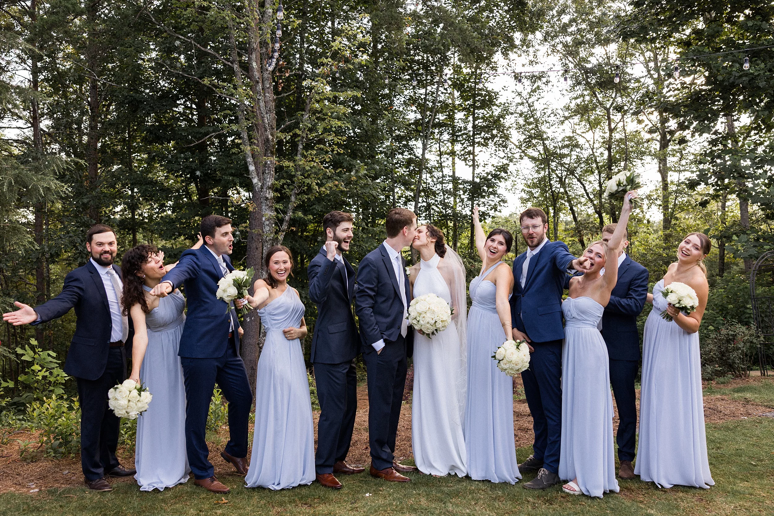 Newlyweds kiss while their wedding party celebrates around them outside in the woods