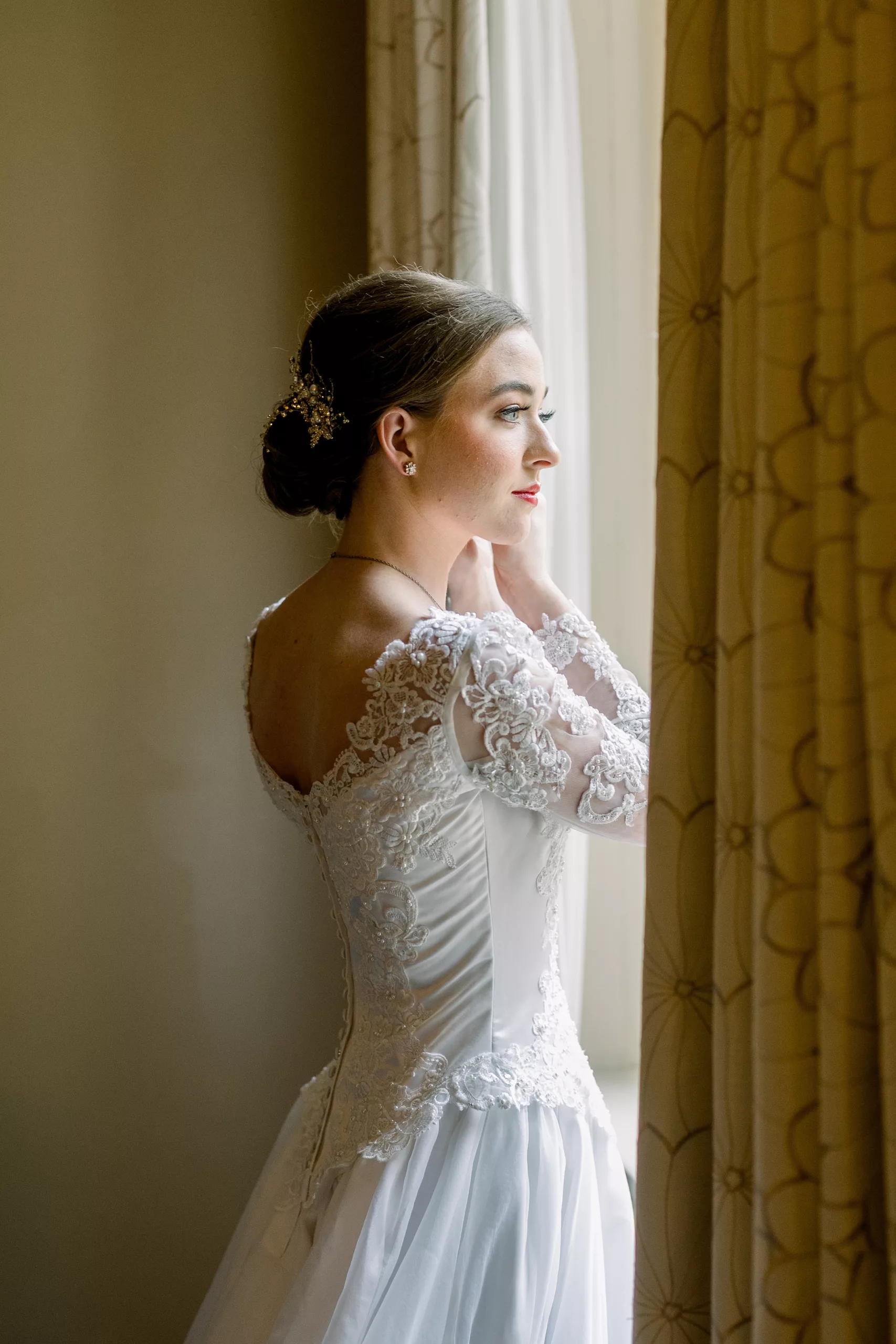 A bride in a white lace dress stands in a window putting on her earrings 