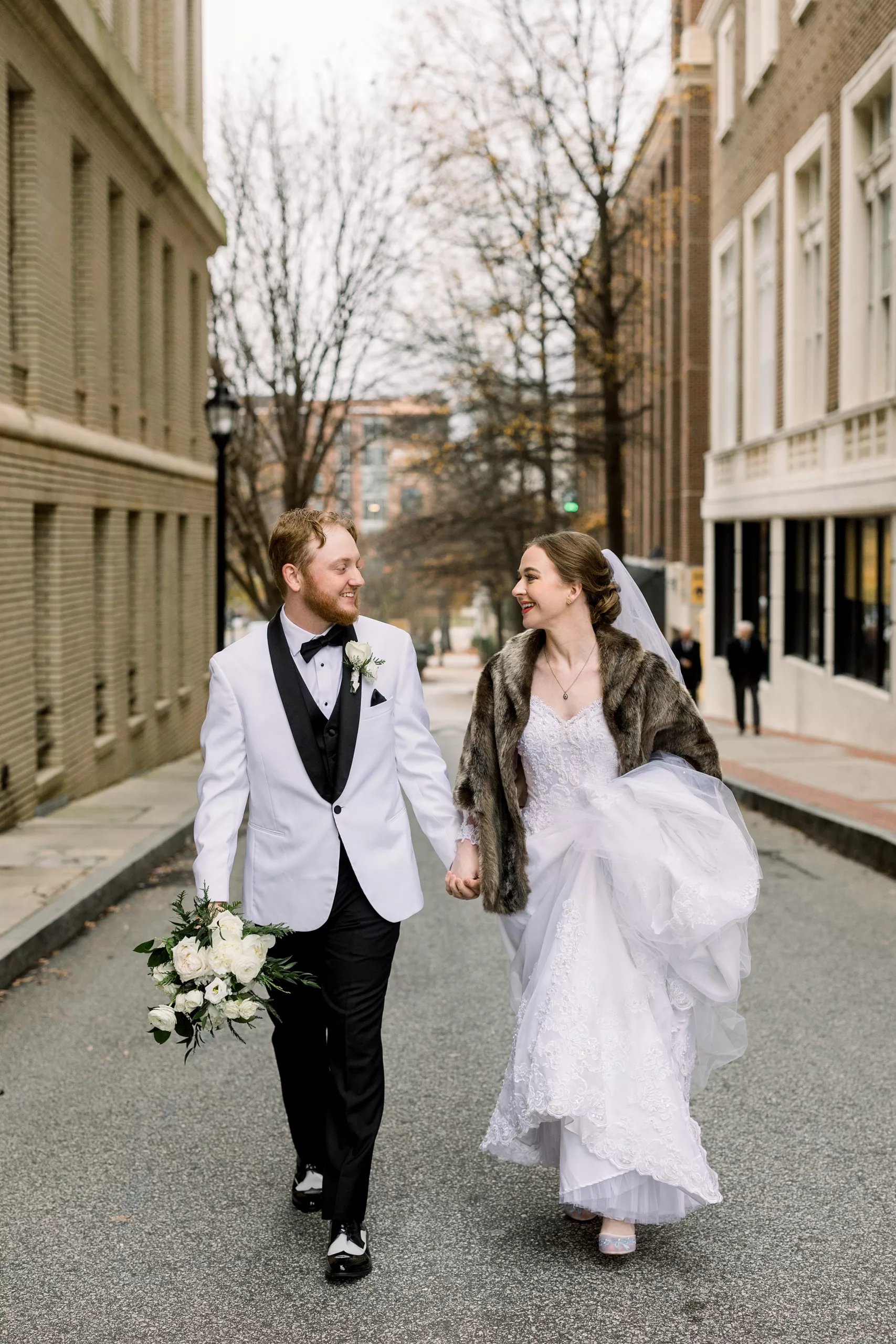 Newlyweds walk down a side street between buildings holding hands while wearing a white suit jacket and fur coat wedding planner vs coordinator