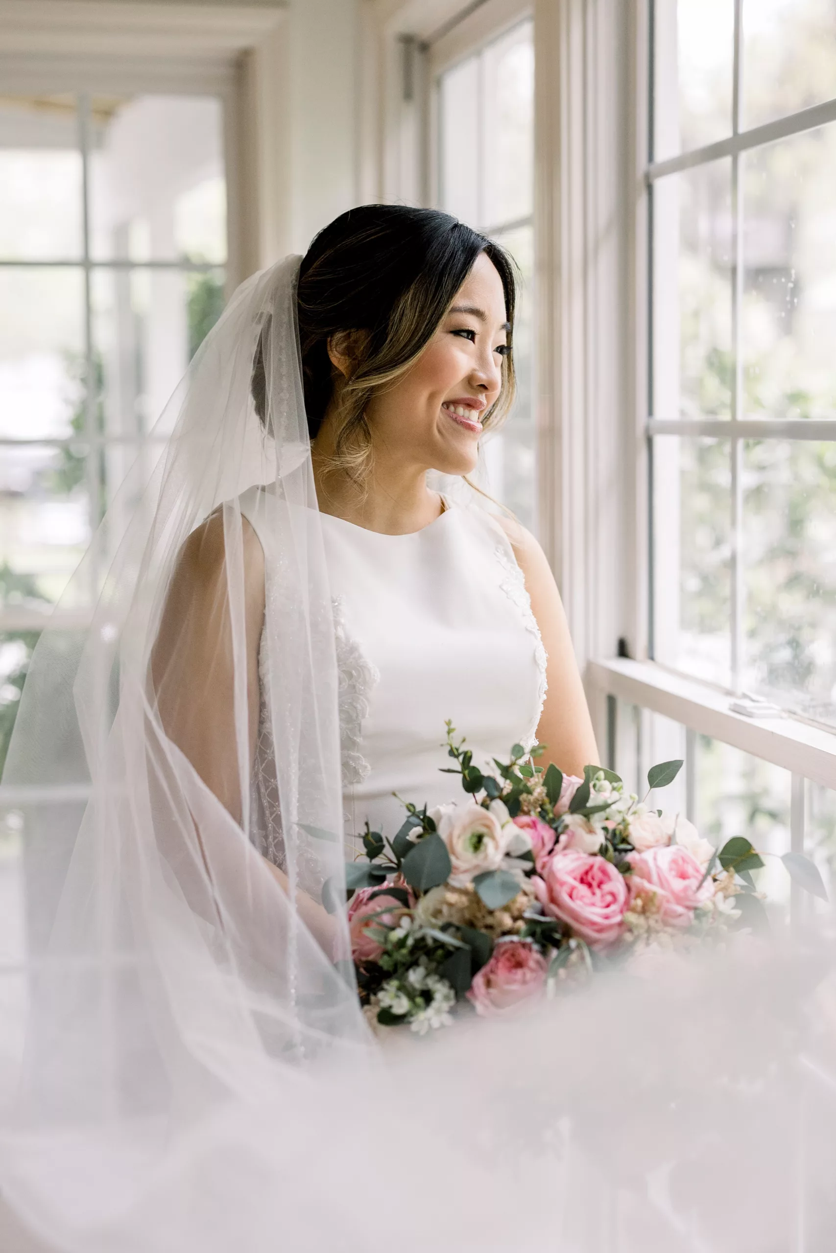 A bride in a silk white dress stands in a sunroom holding her pink bouquet wedding planner vs coordinator