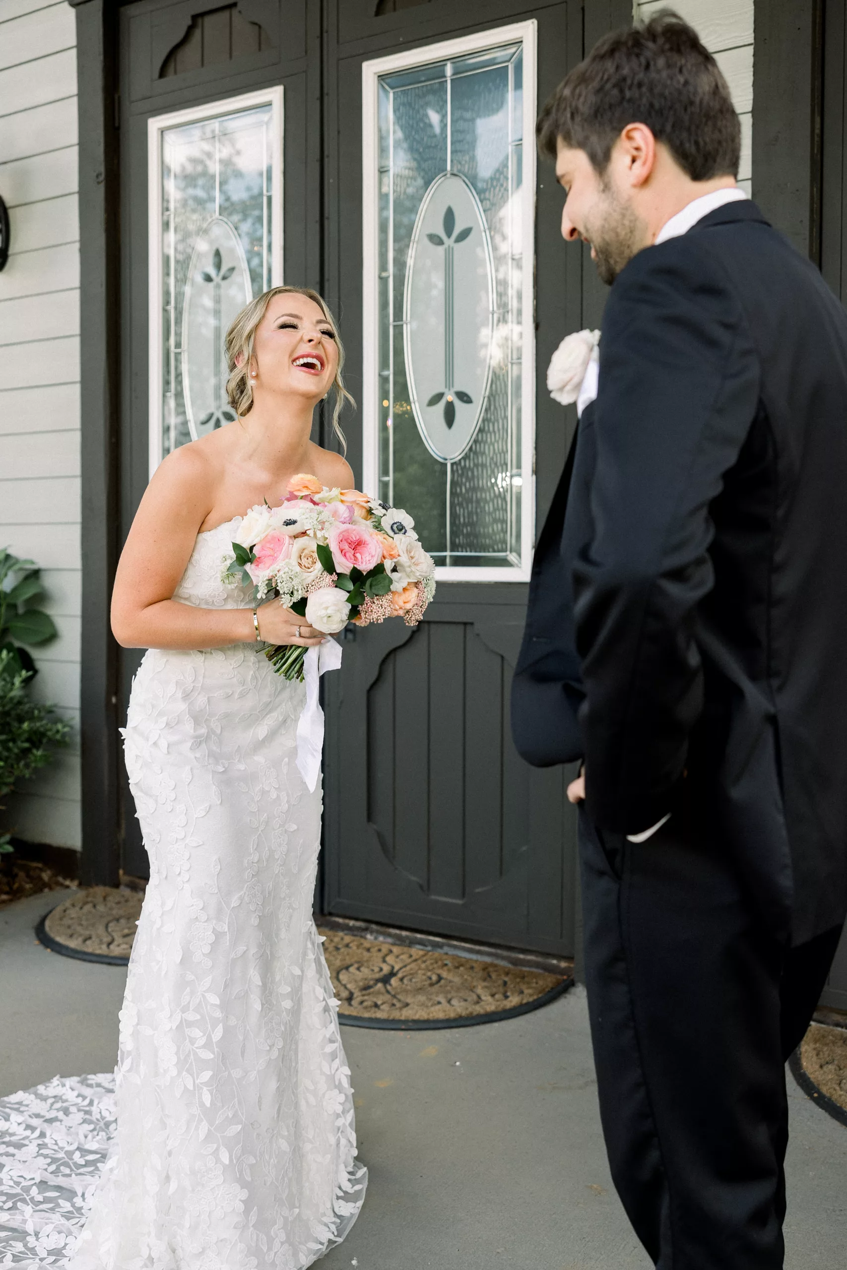A bride laughs in her dress while holding her pink bouquet as her groom in a black suit sees her for the first time before the ceremony