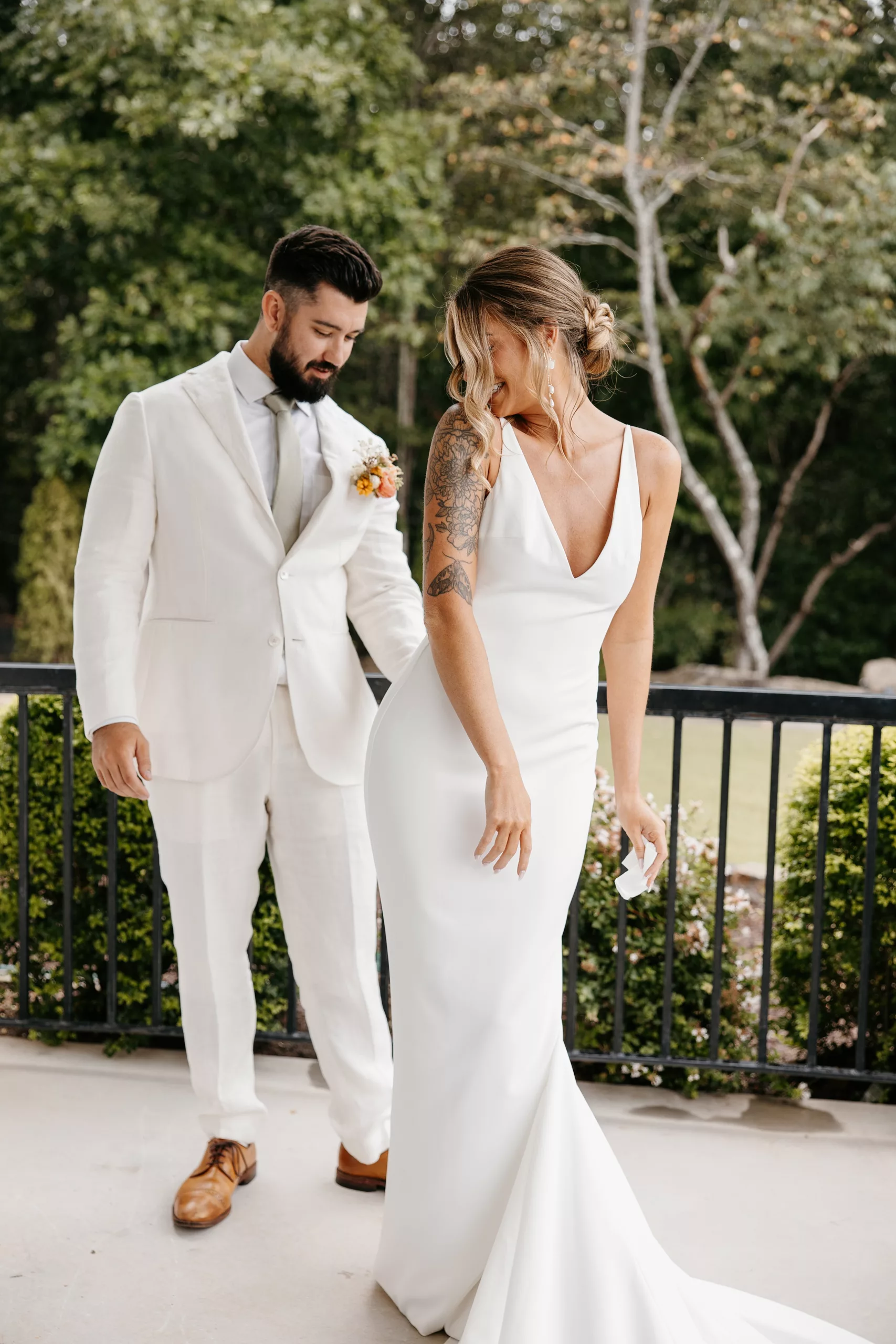 A groom looks his bride up and down in a white suit and her silk dress on a patio as he sees her for the first time