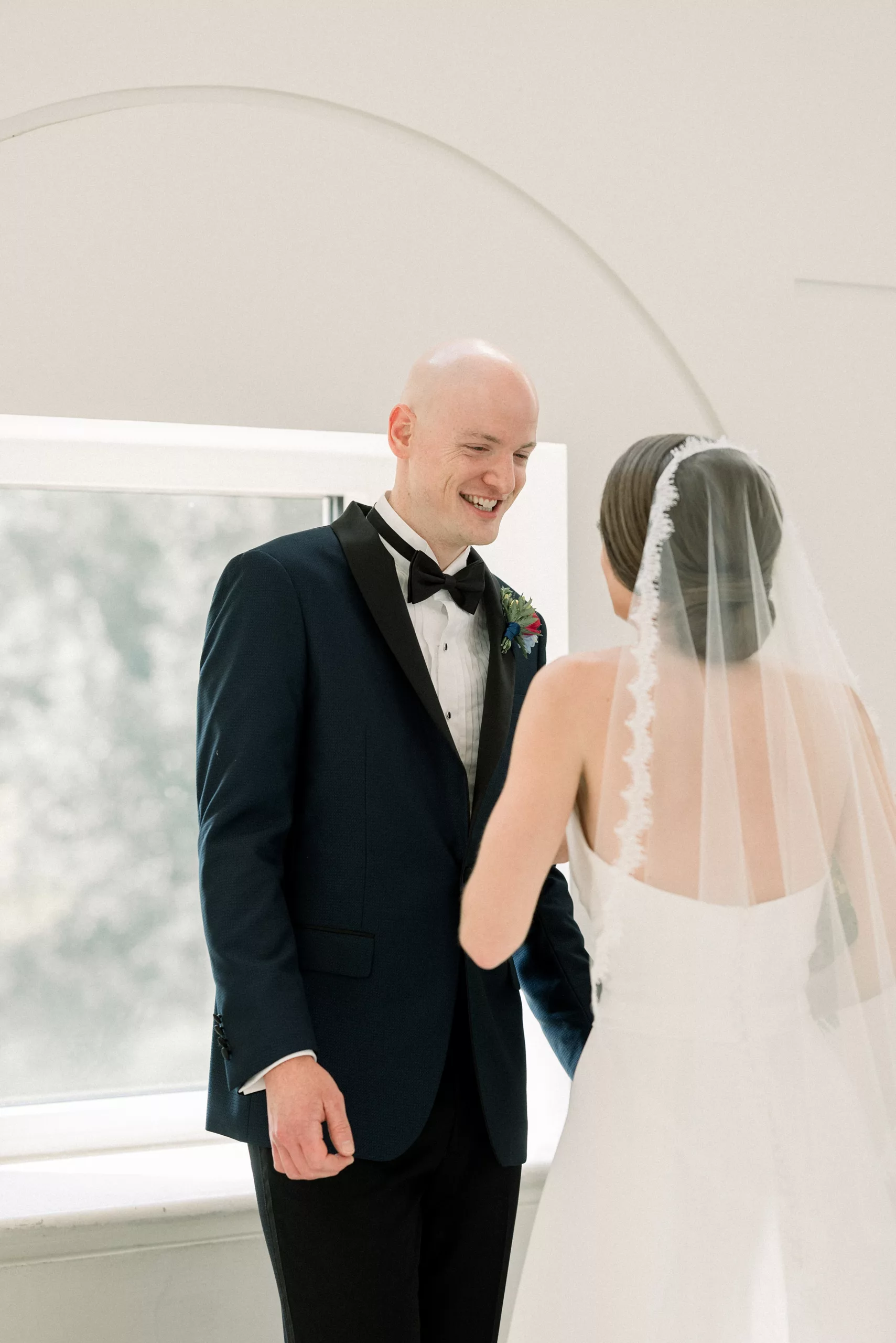 A groom smiles big as he sees his bride in her dress in a white room in front of a window