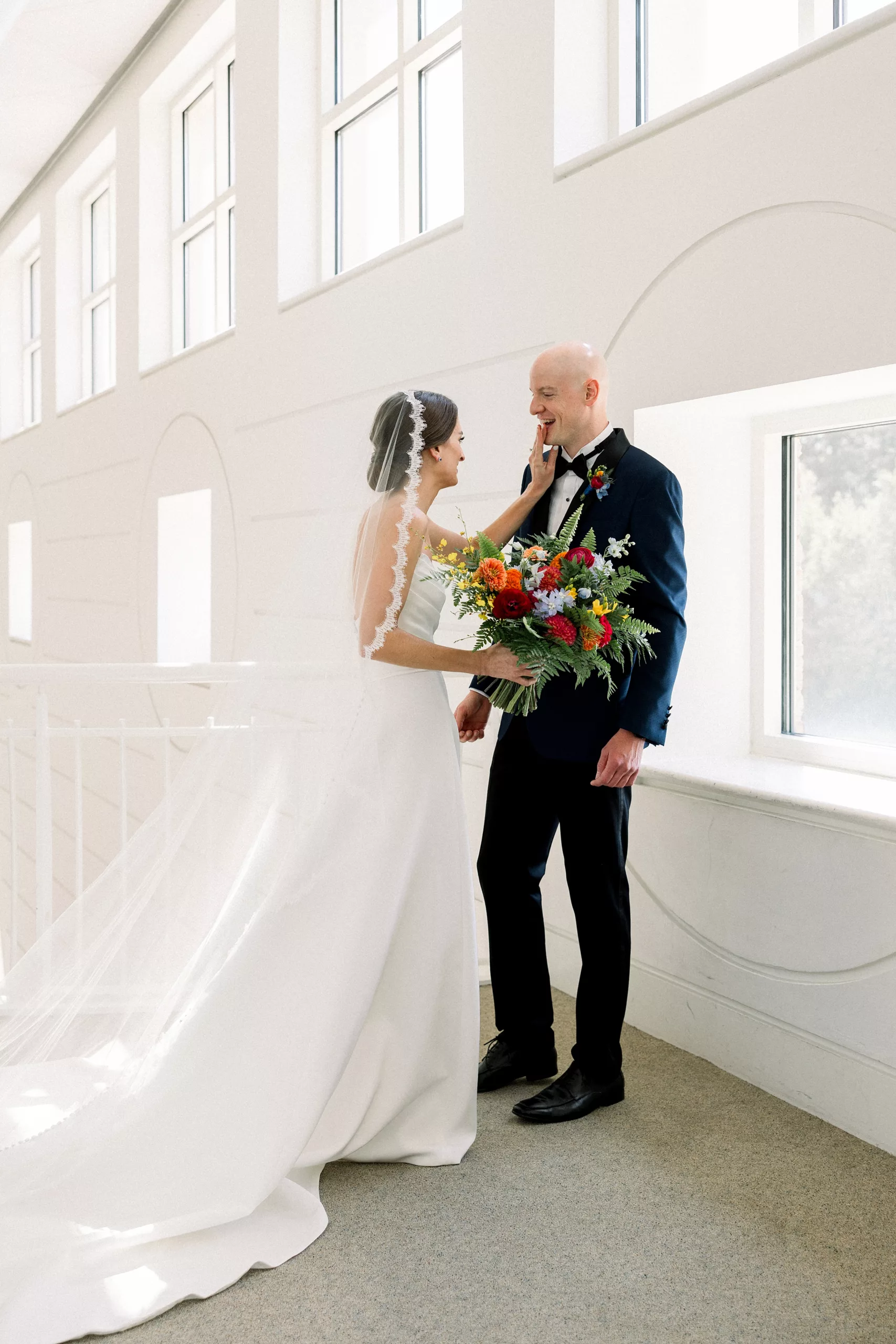 A bride pushes her groom's chin back up as he sees her for the first time in her dress in a white building