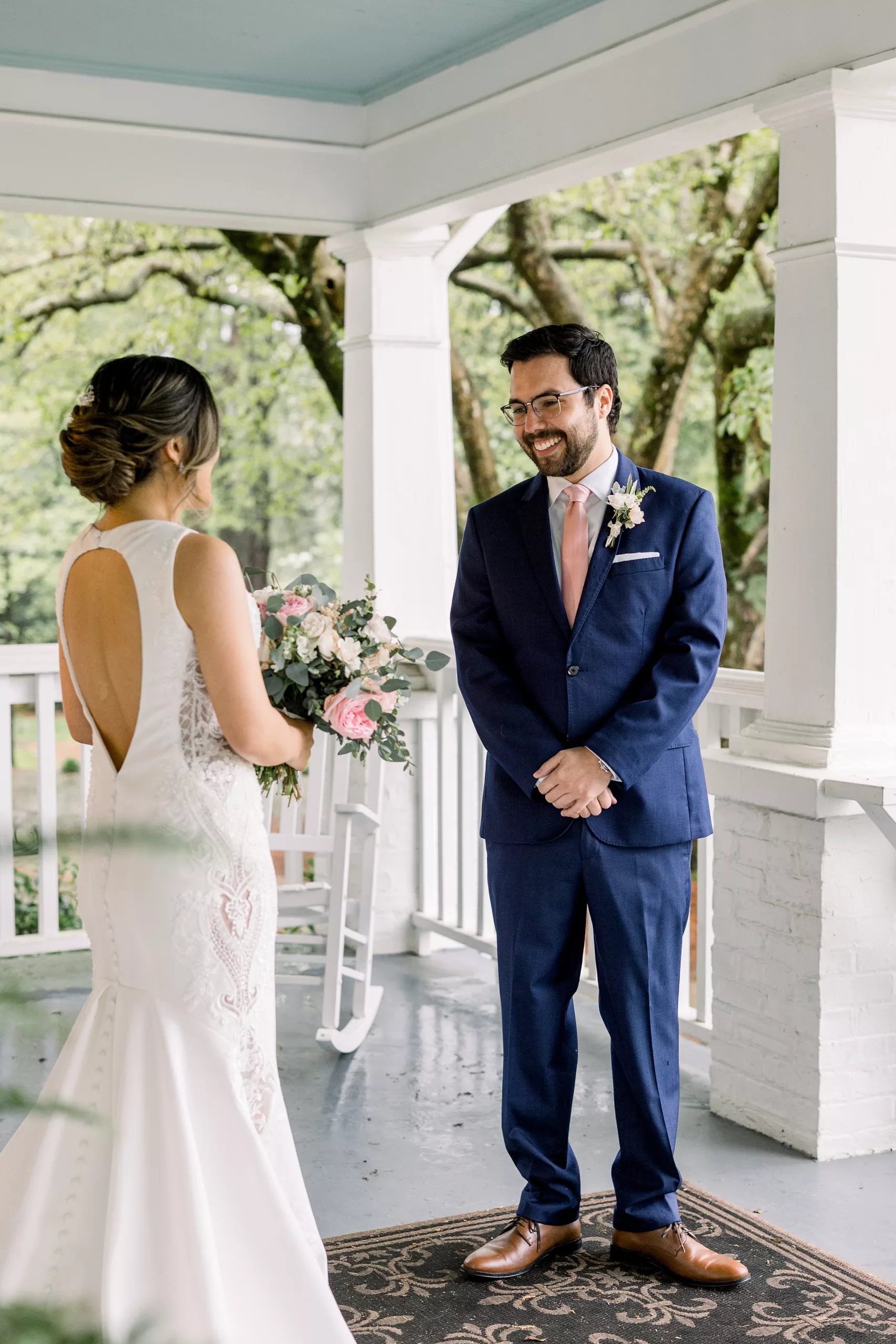 A groom stands with hands crossed smiling at his bride as he sees her for the first time on a porch during their first look wedding