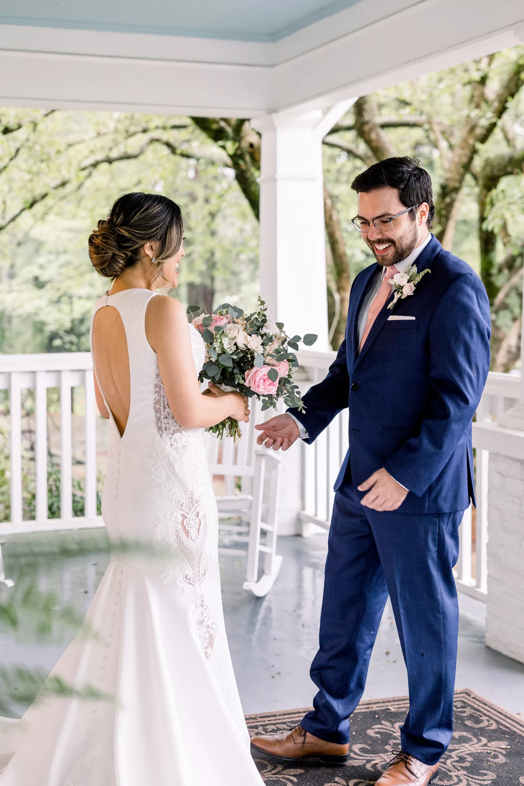A groom smiles at his bride while wearing a blue suit and pink tie as he sees her for the first time in her dress during a pre wedding first look