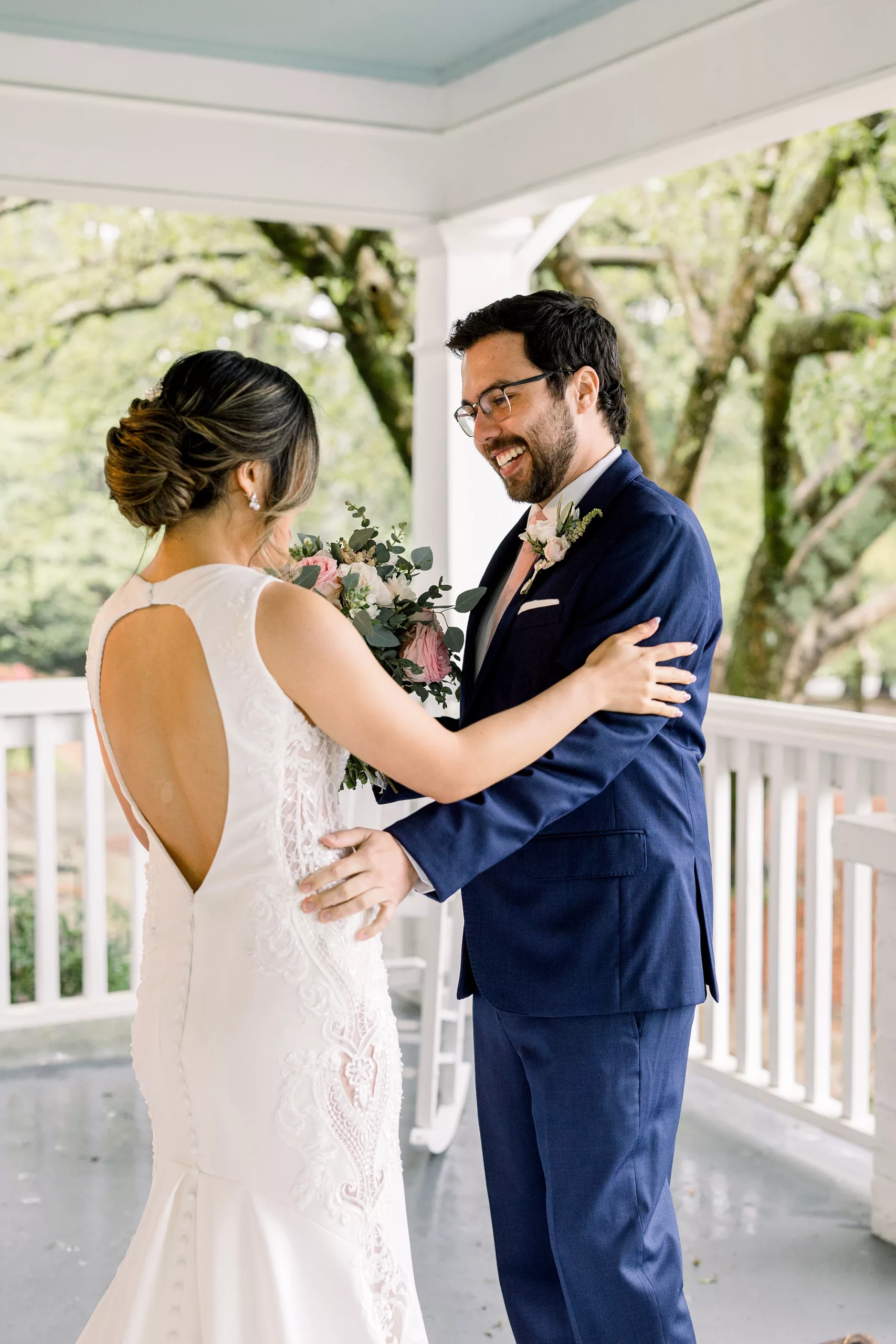 A bride and groom lean in for a big hug while smiling and standing on a porch in a blue suit and white lace embroidered dress during their first look wedding