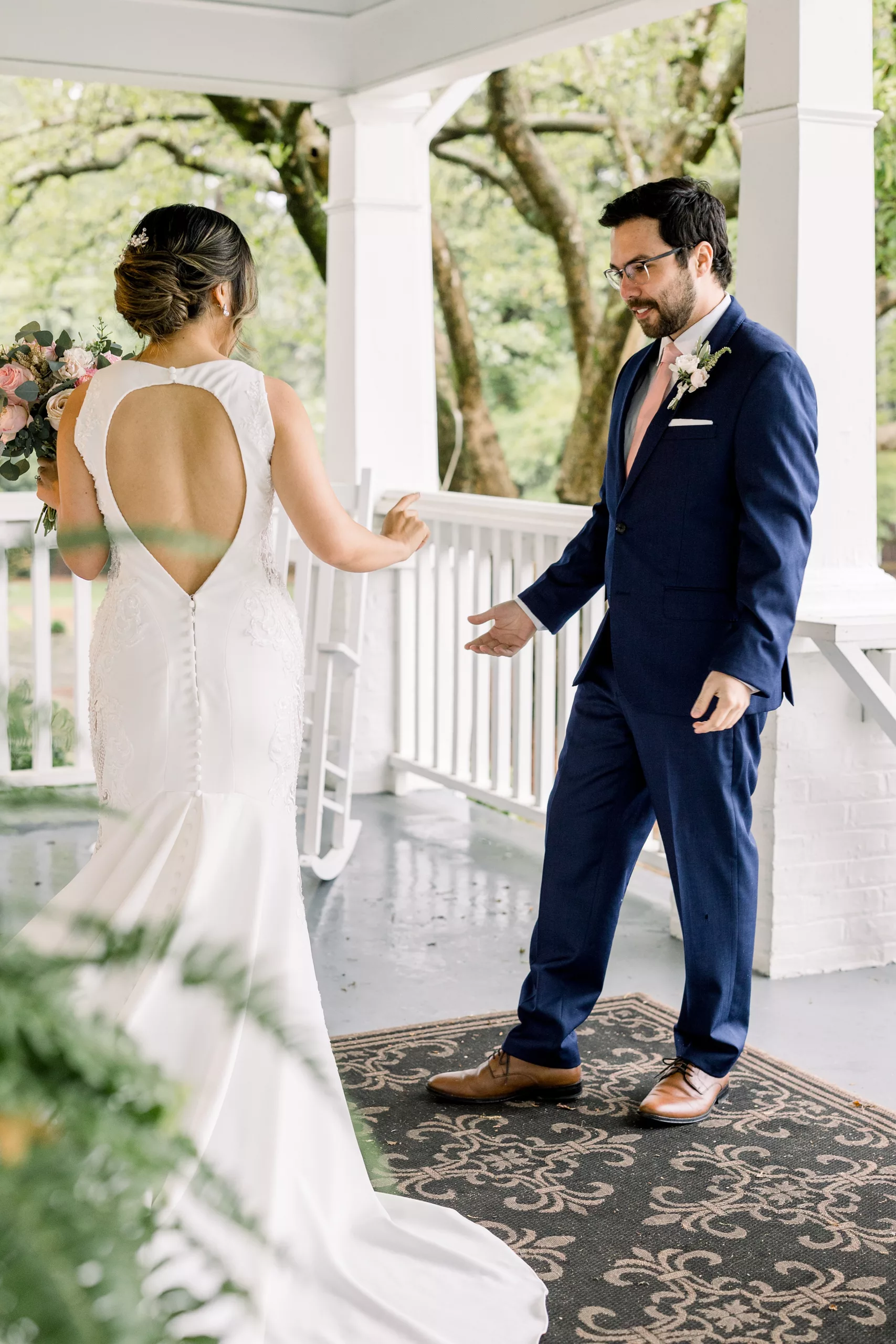 A groom in a blue suit and pink tie looks on in amazement as his bride twirls during their first look before the wedding ceremony
