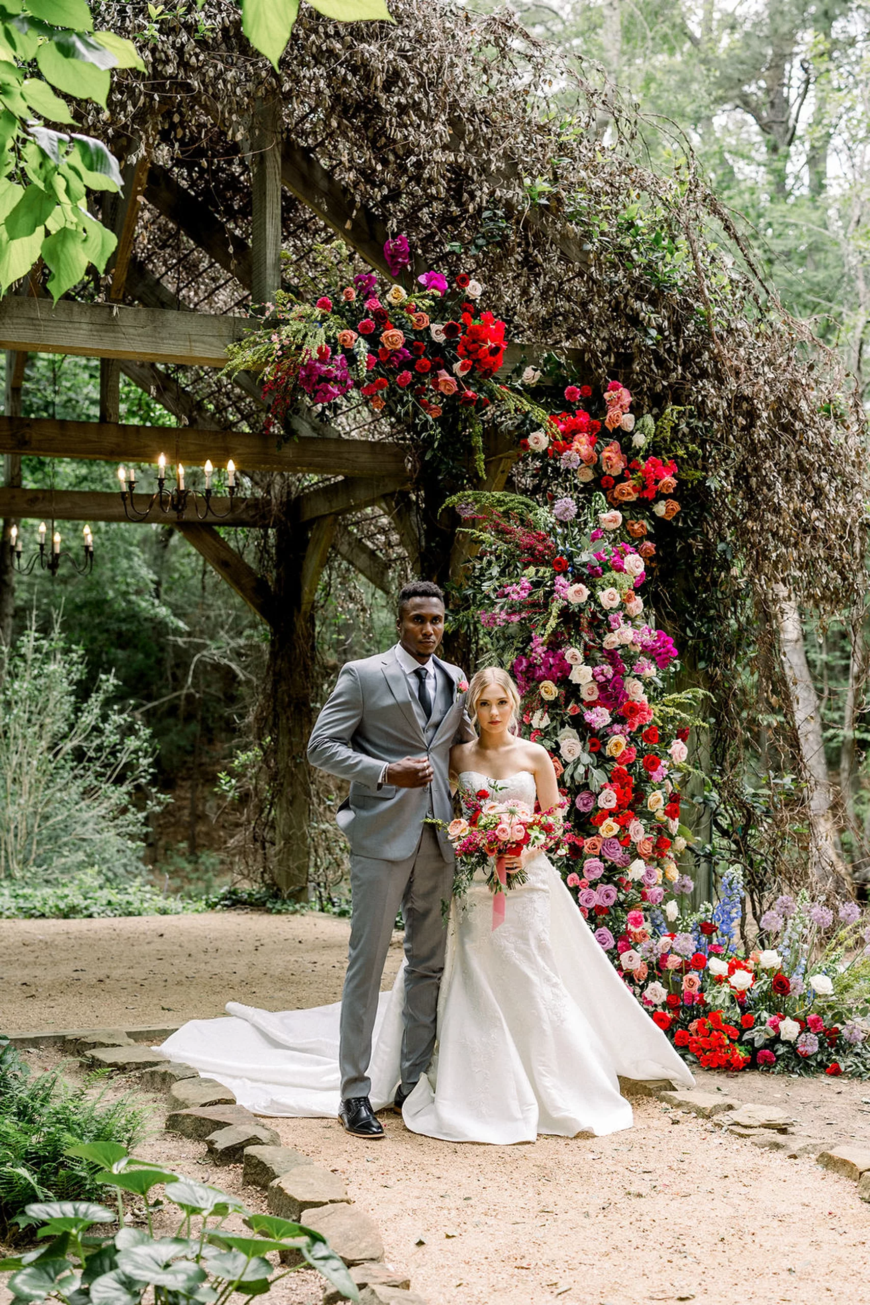 A groom in a grey suit walks his bride down a gravel path in a garden away from a rose covered pergola at the iron manor wedding venue
