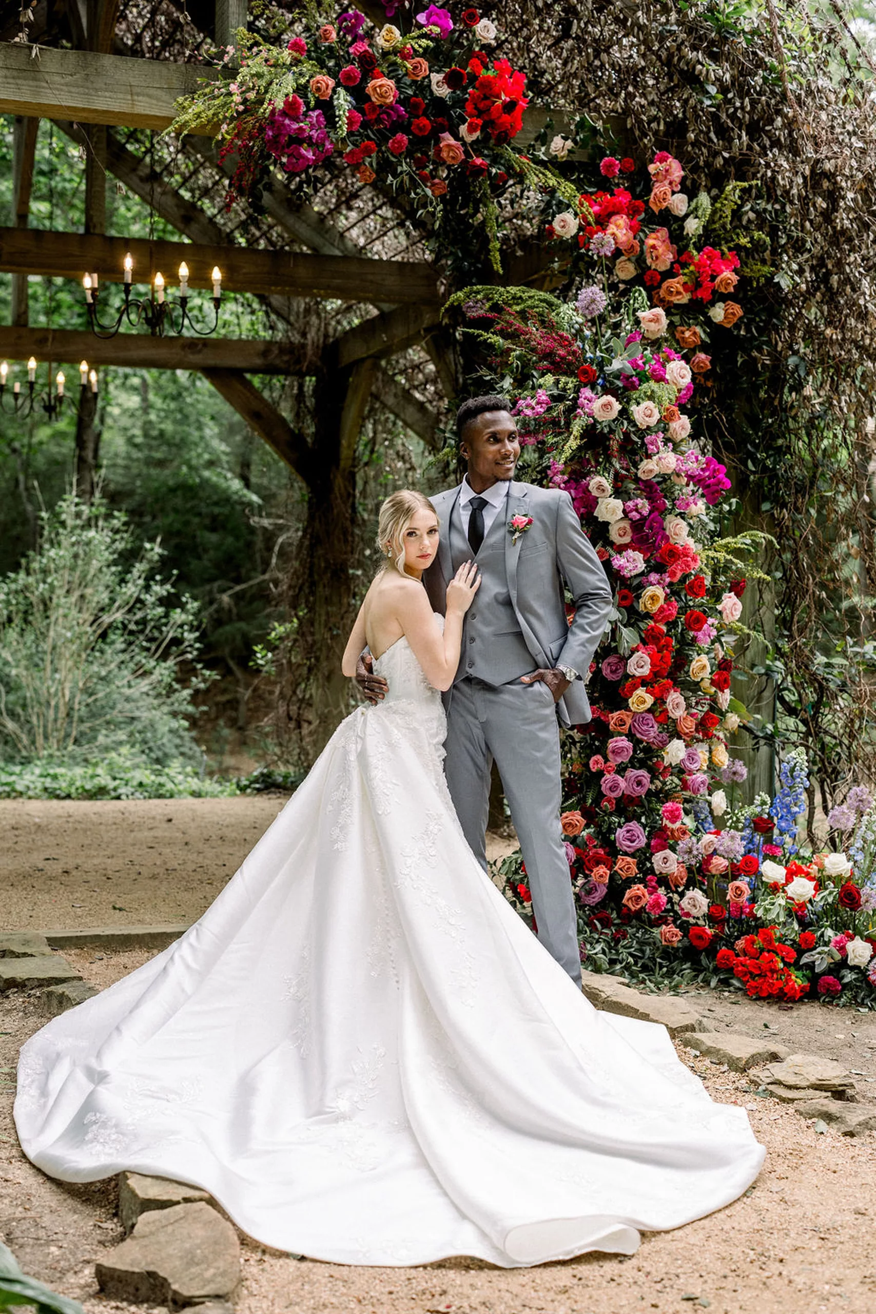 A groom stands under a wall of roses with his bride leaning onto him with her hand on his chest and her train spread over the gravel path at the iron manor wedding venue