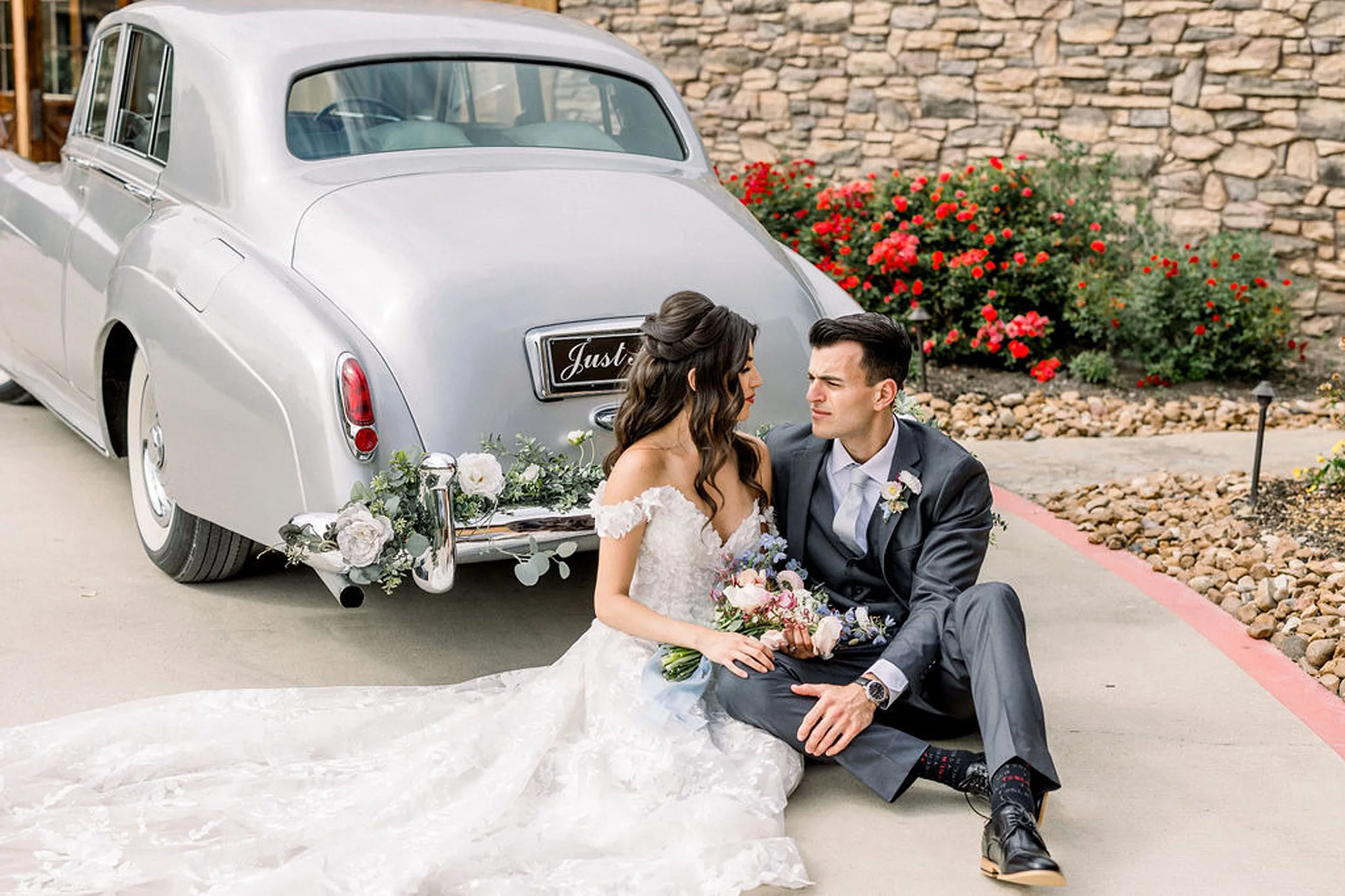 Newlyweds sit behind a silver antique car in the iron manor wedding venue driveway surrounded by red roses