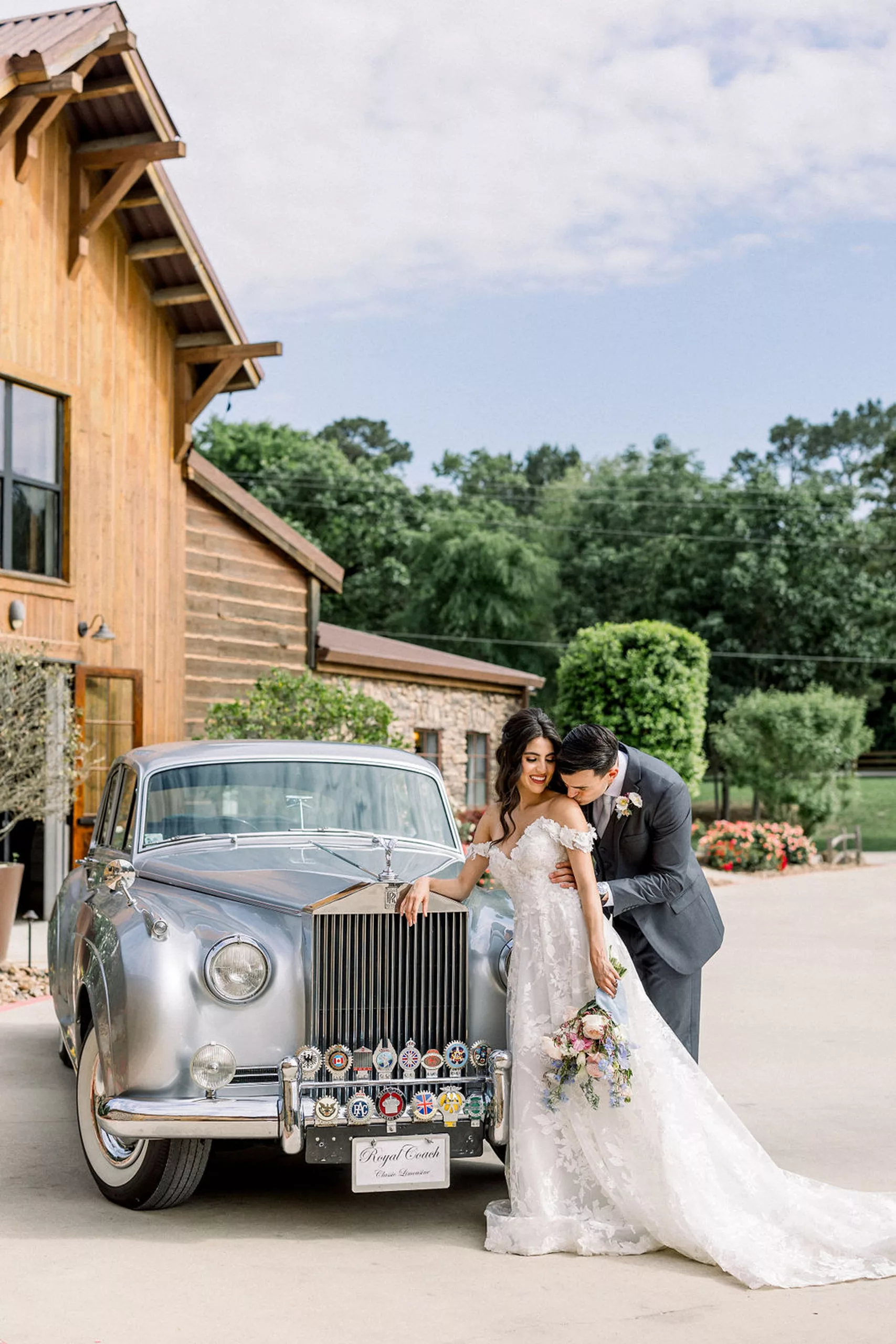 A groom in a grey suit kisses the shoulder of his bride as she leans on a silver Rolls Royce in the driveway of the iron manor wedding venue