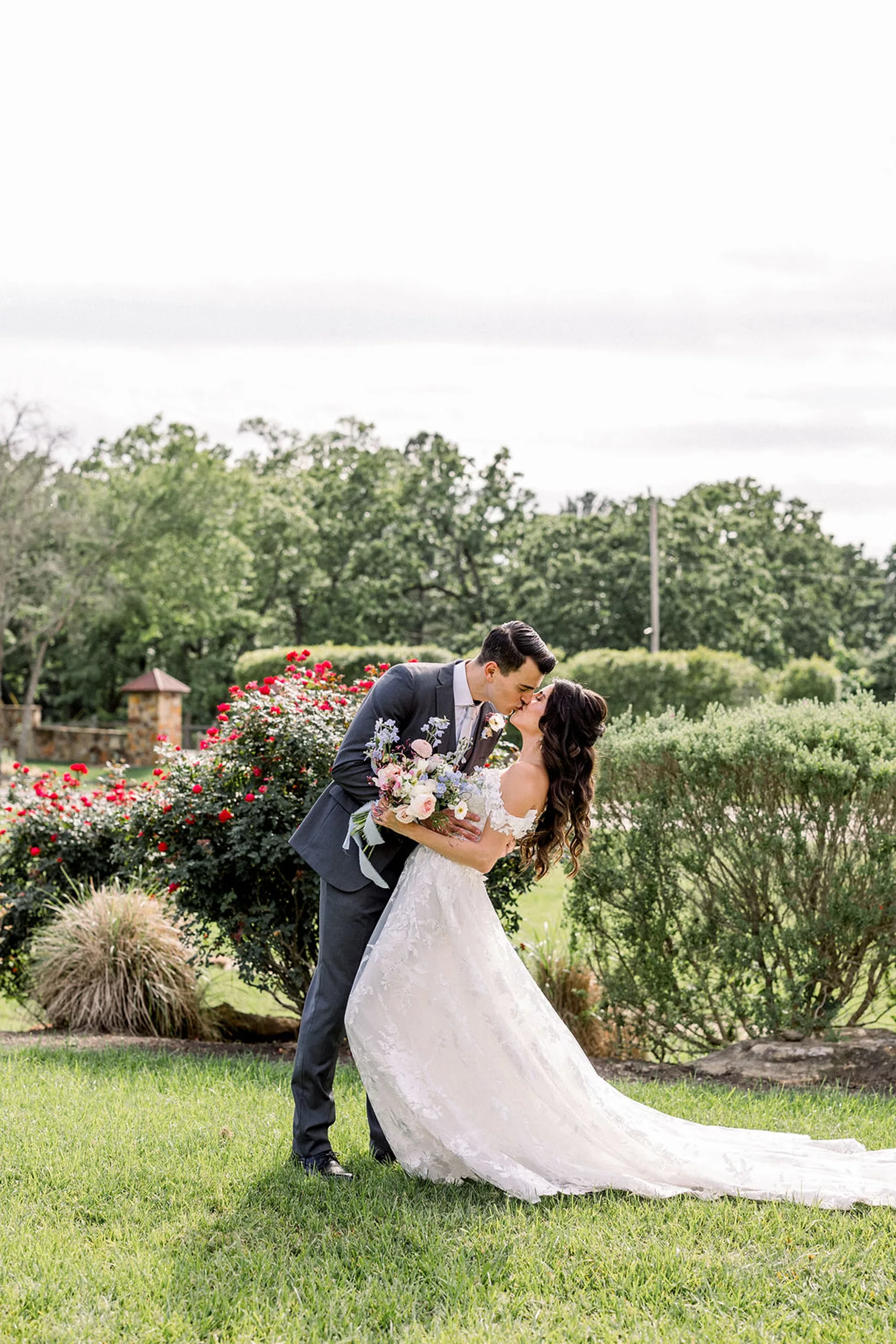 A groom dips and kisses his bride in a white lace dress in the iron manor wedding venue rose garden