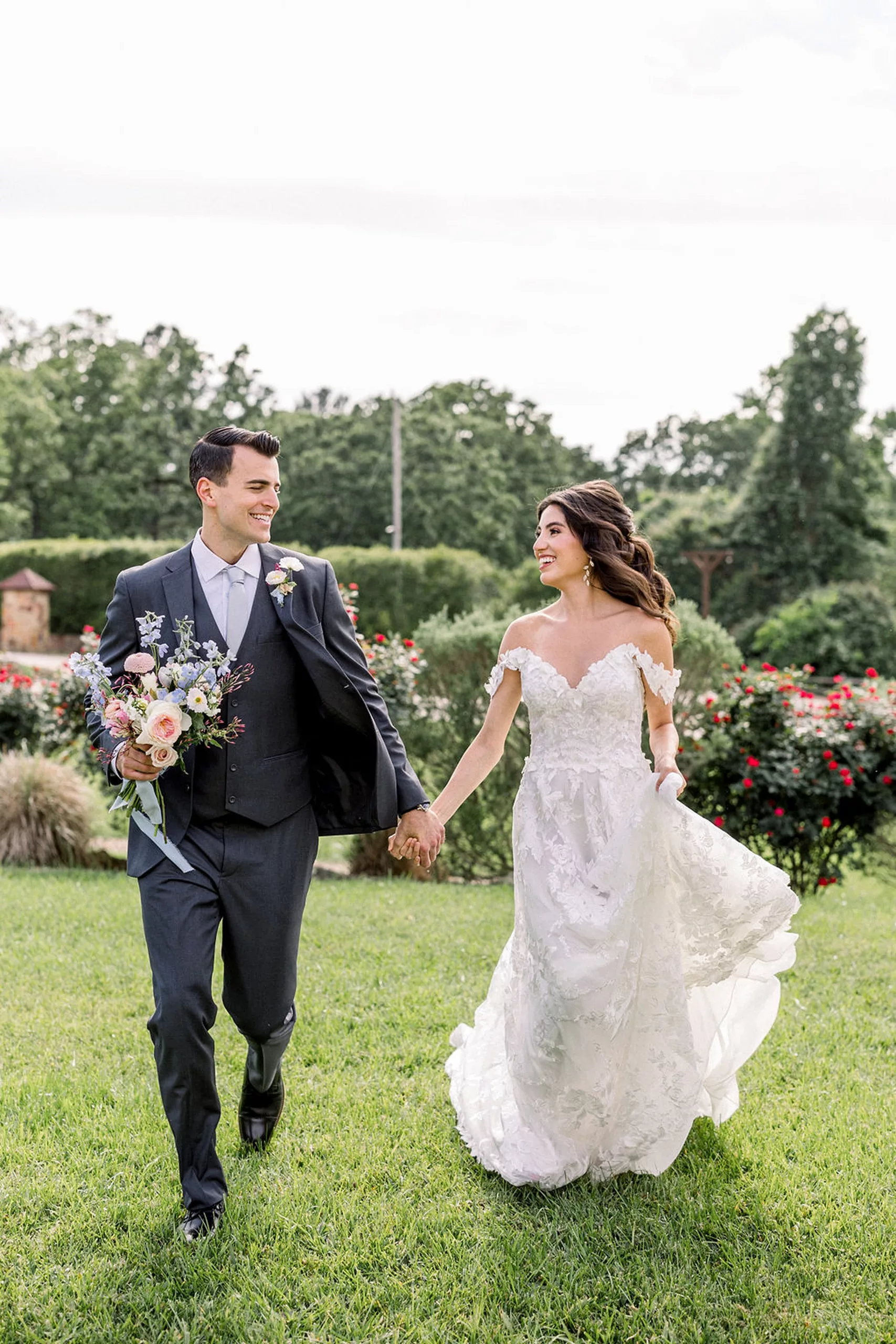 A bride and groom walk hand in hand through a rose garden at an iron manor wedding as the groom in a grey suit holds the bouquet
