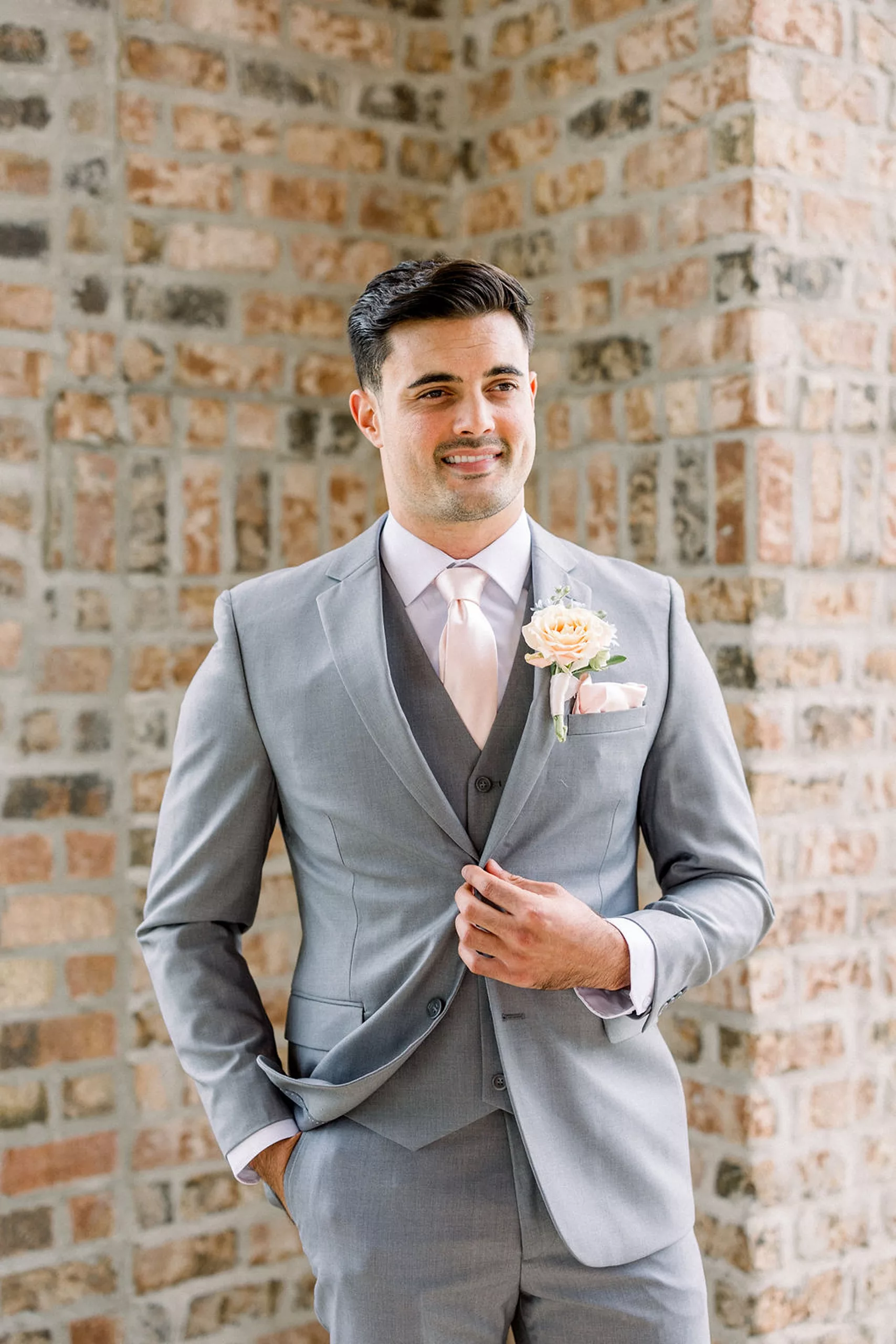 A groom in a grey suit and pink tie stands in a patio buttoning his suit jacket