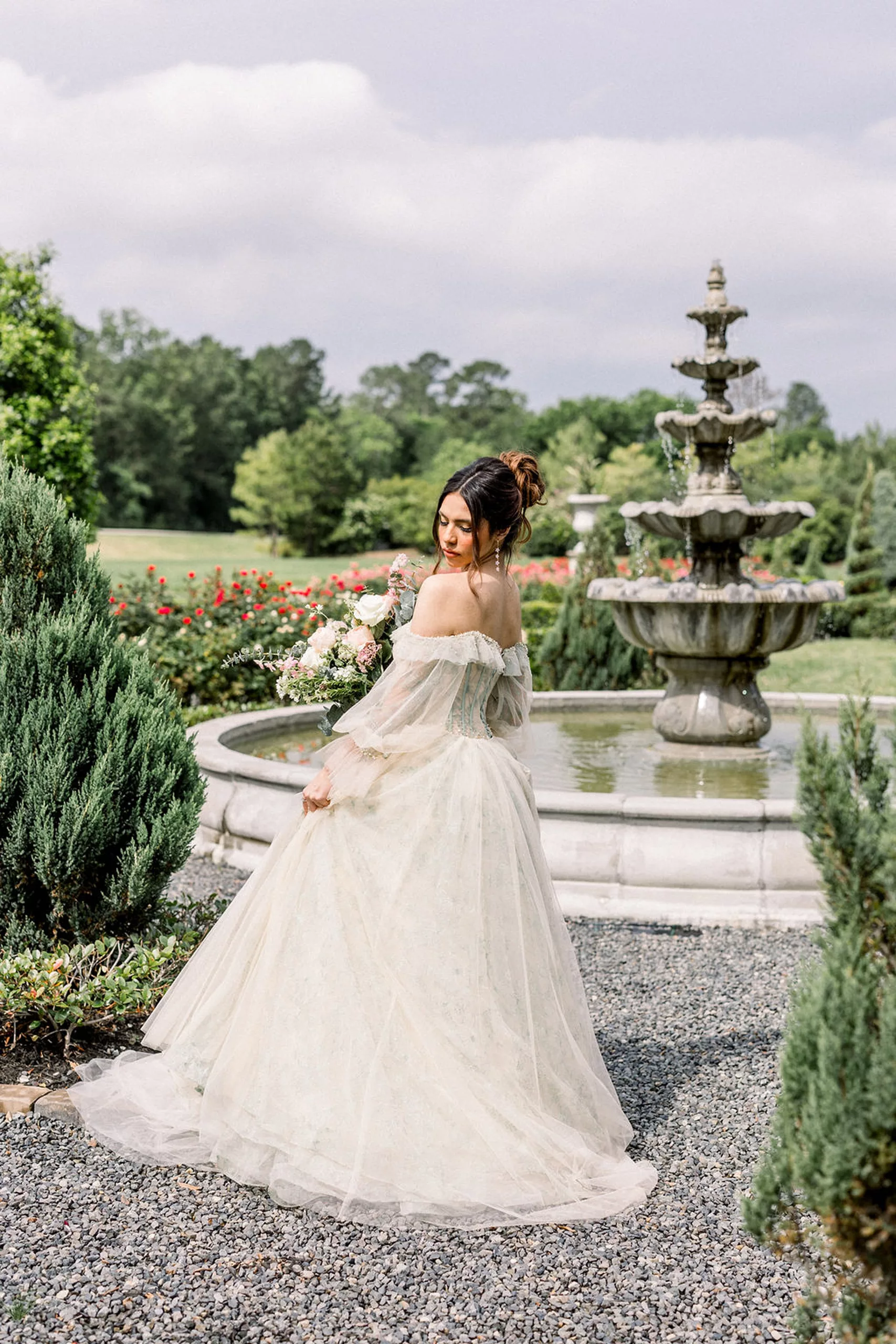 A bride twirls and looks down at her train while standing in a gravel path by a garden fountain