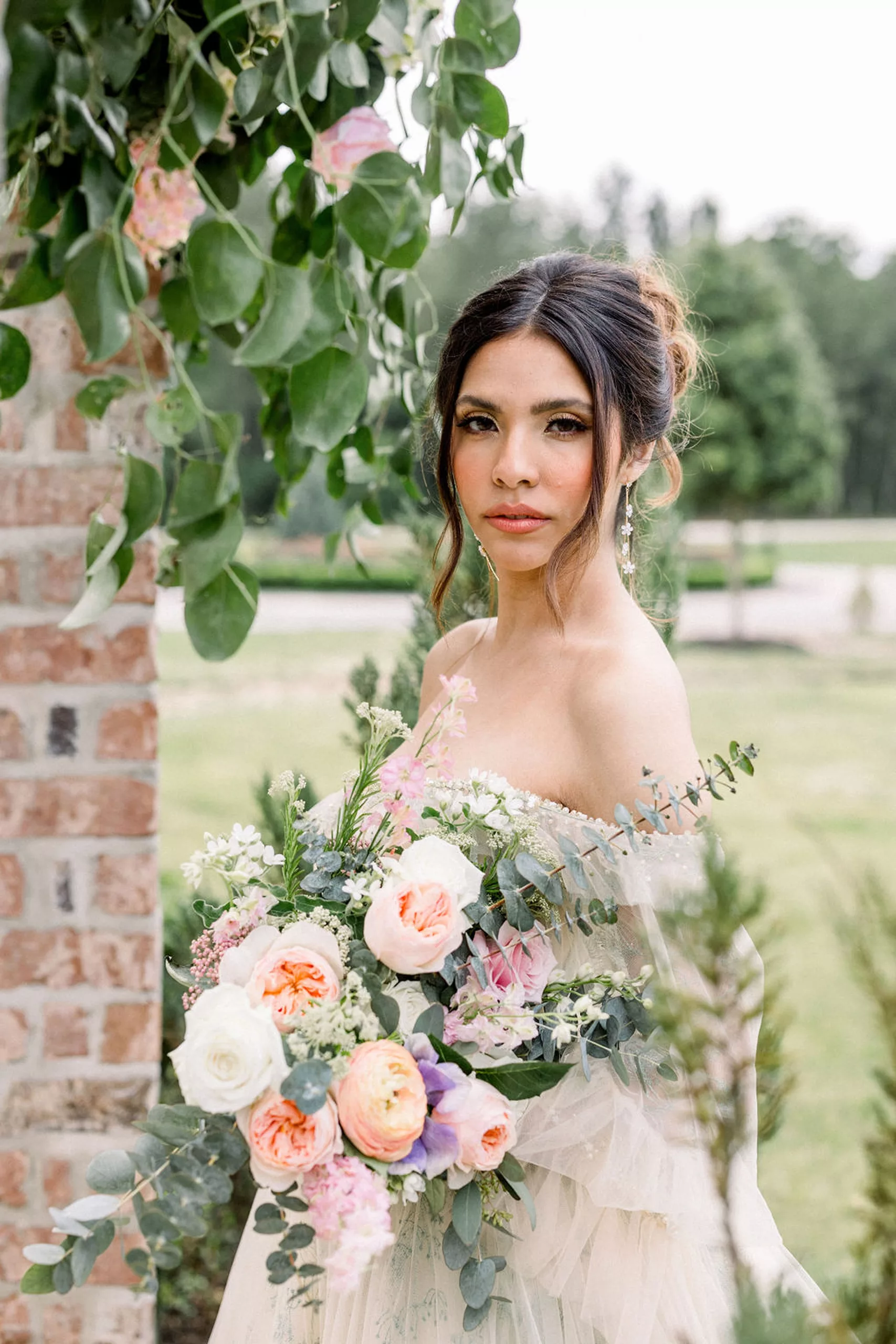 A bride stands in a garden holding a large pink and white rose bouquet and wearing a velour dress