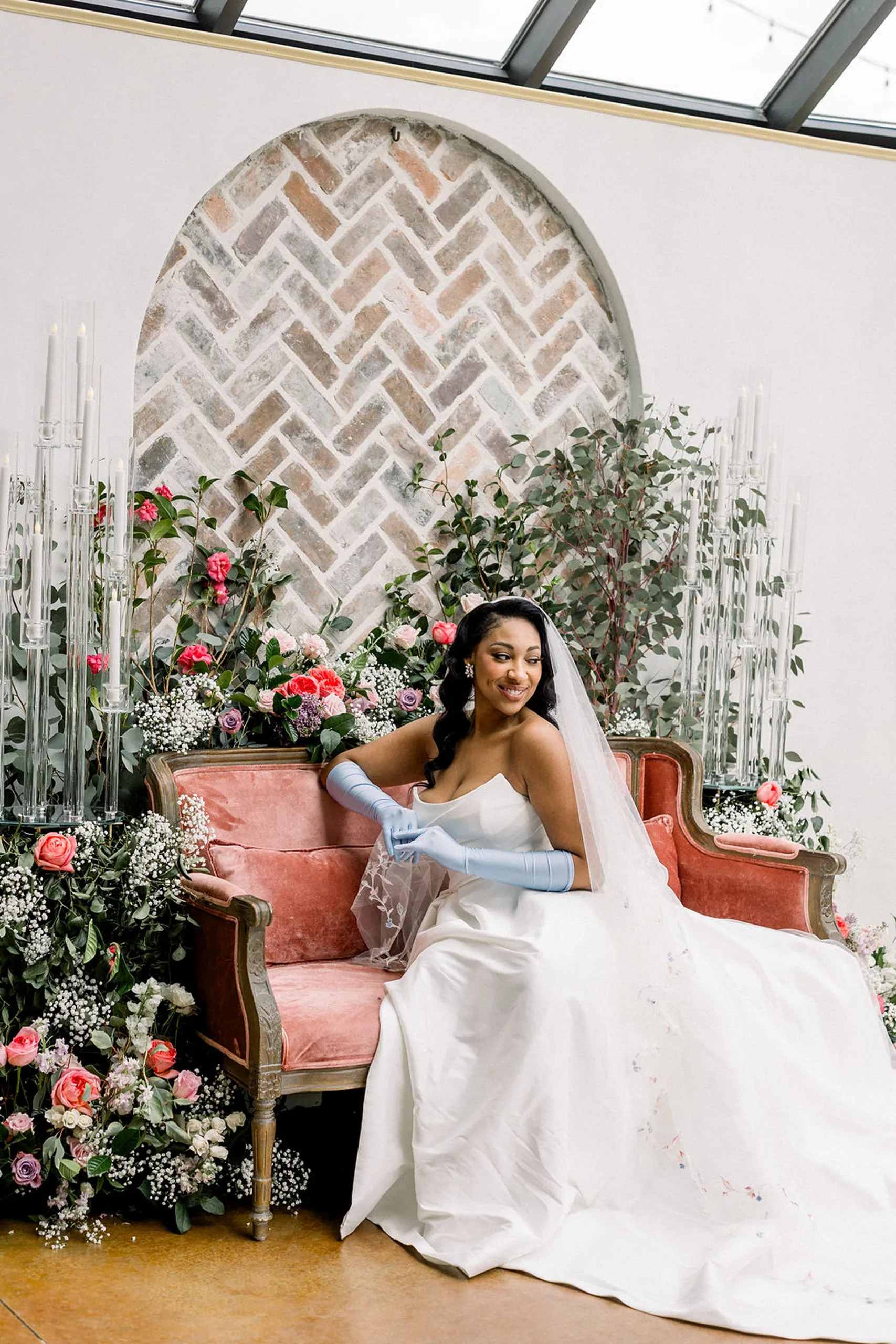 A bride smiles down her shoulder while sitting on a pink couch surrounded by flowers and candles