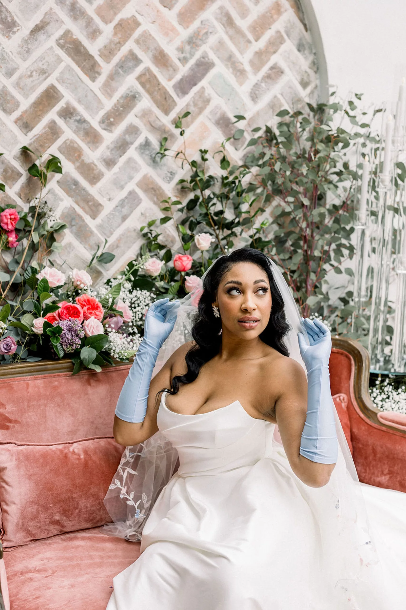 A bride plays with her veil while wearing blue gloves while sitting on a pink couch surrounded by flowers