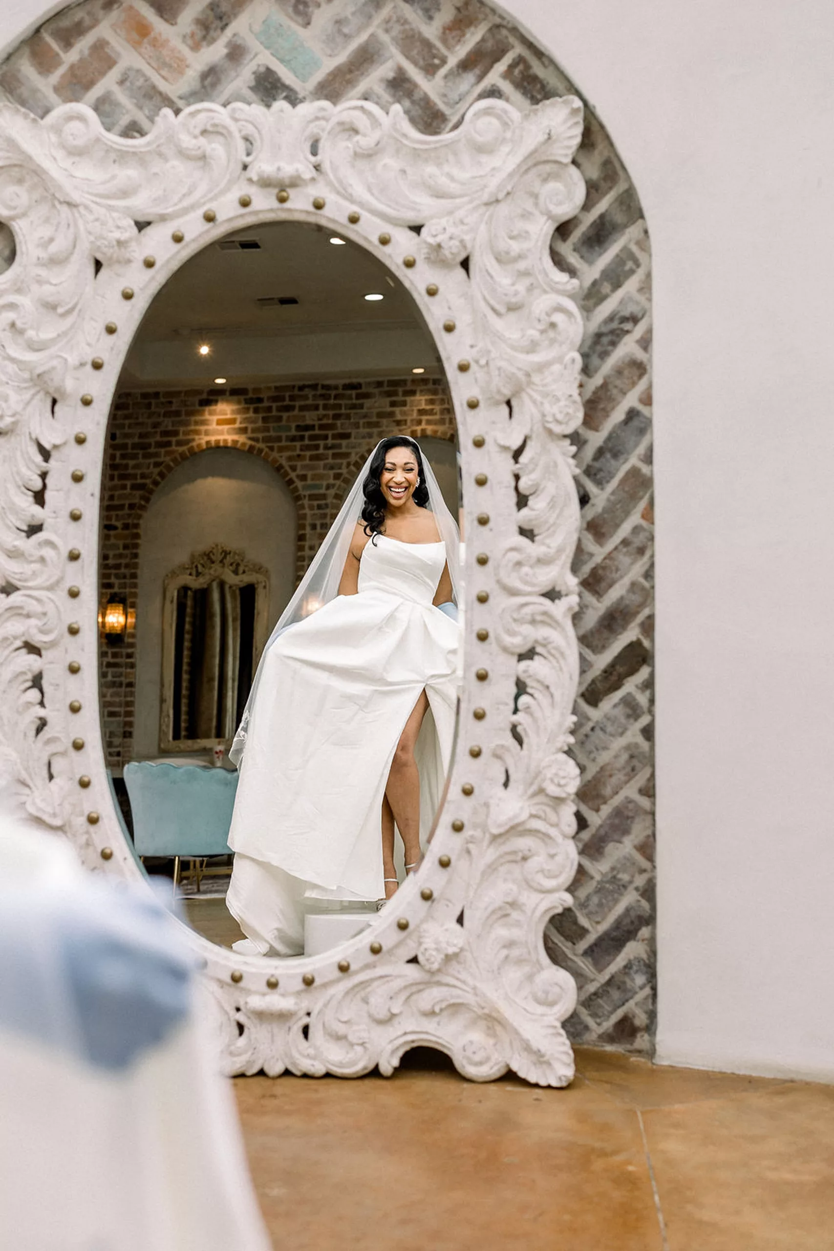 A bride sees herself in her dress in a large ornate mirror in the getting ready room