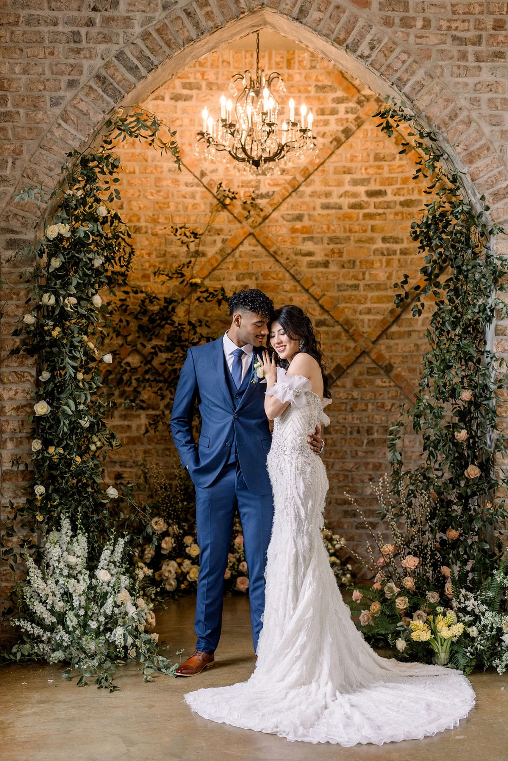 A groom stands under an arch decorated with lots of flowers holding his bride close under a chandelier