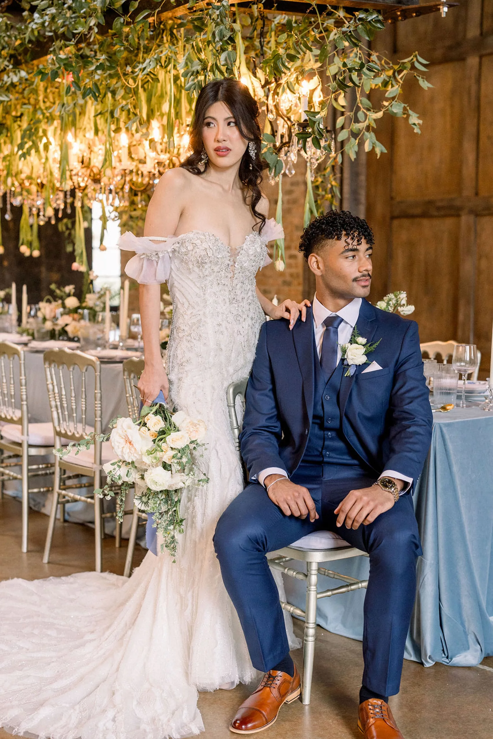 A groom in a blue suit sits at a wedding reception table with silver chairs and blue linens with his bride standing behind him with a hand on his shoulder