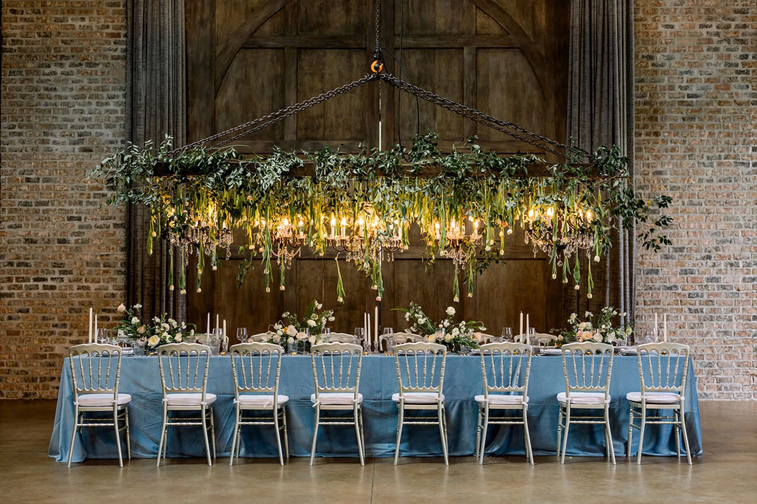 Details of a blue wedding reception table with silver chairs in front of a large wooden door under a large chandelier dressed with hanging tulips and chandeliers