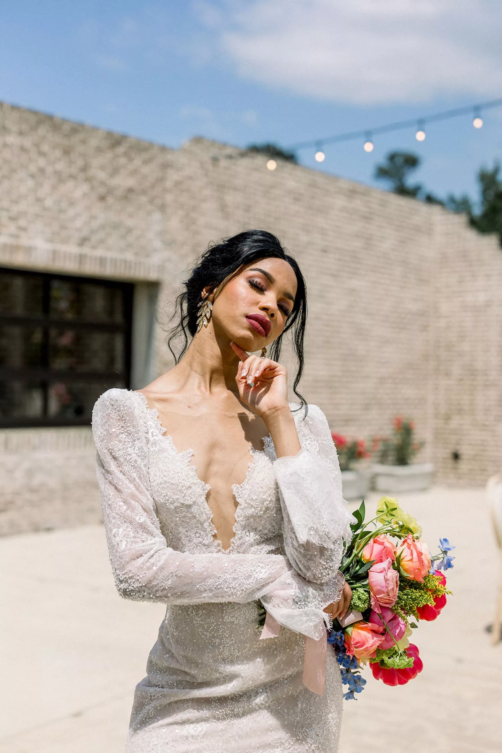 A bride in a white lace dress stands in the sun on a patio while holding a finger under her chin and her bouquet in the other hand at a iron manor wedding