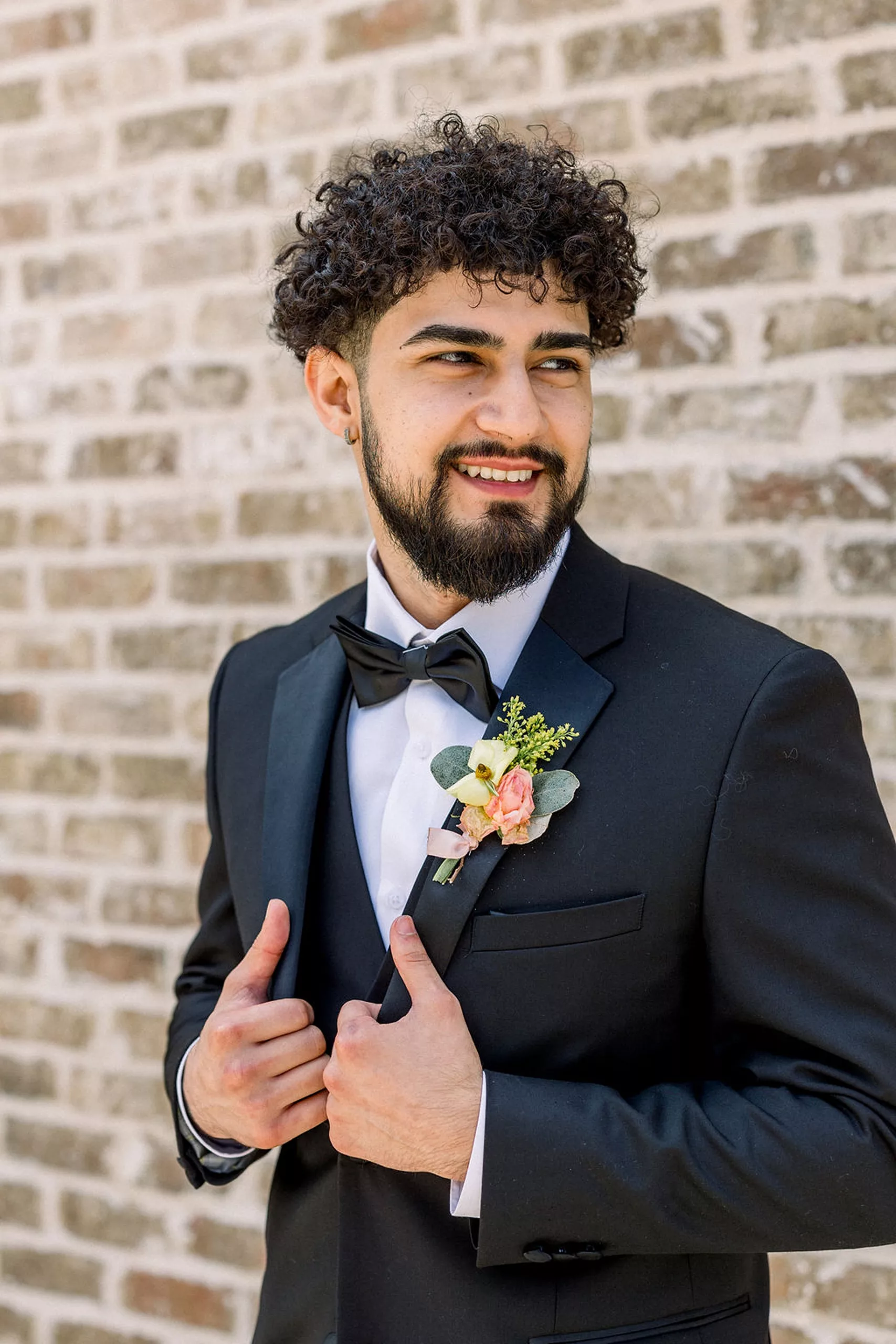 A groom in a black tuxedo and floral boutonniere stands in front of a brick wall holding his lapels and looking over his shoulders