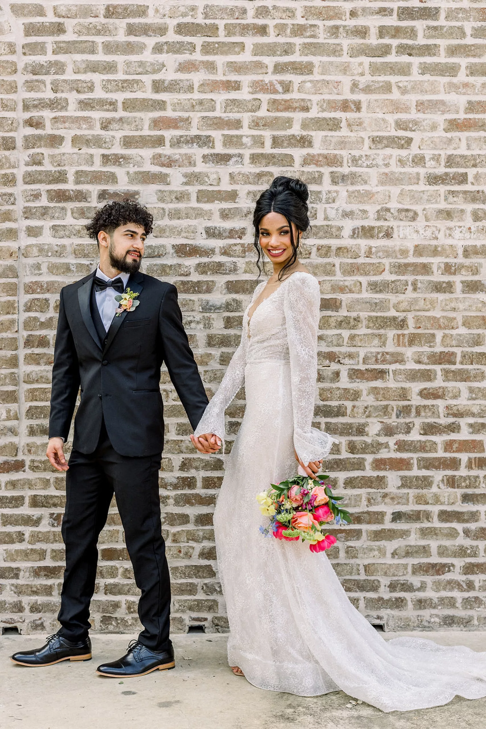 A groom in a black tuxedo leads his bride in a lace dress by the hand around a brick wall at the iron manor wedding venue