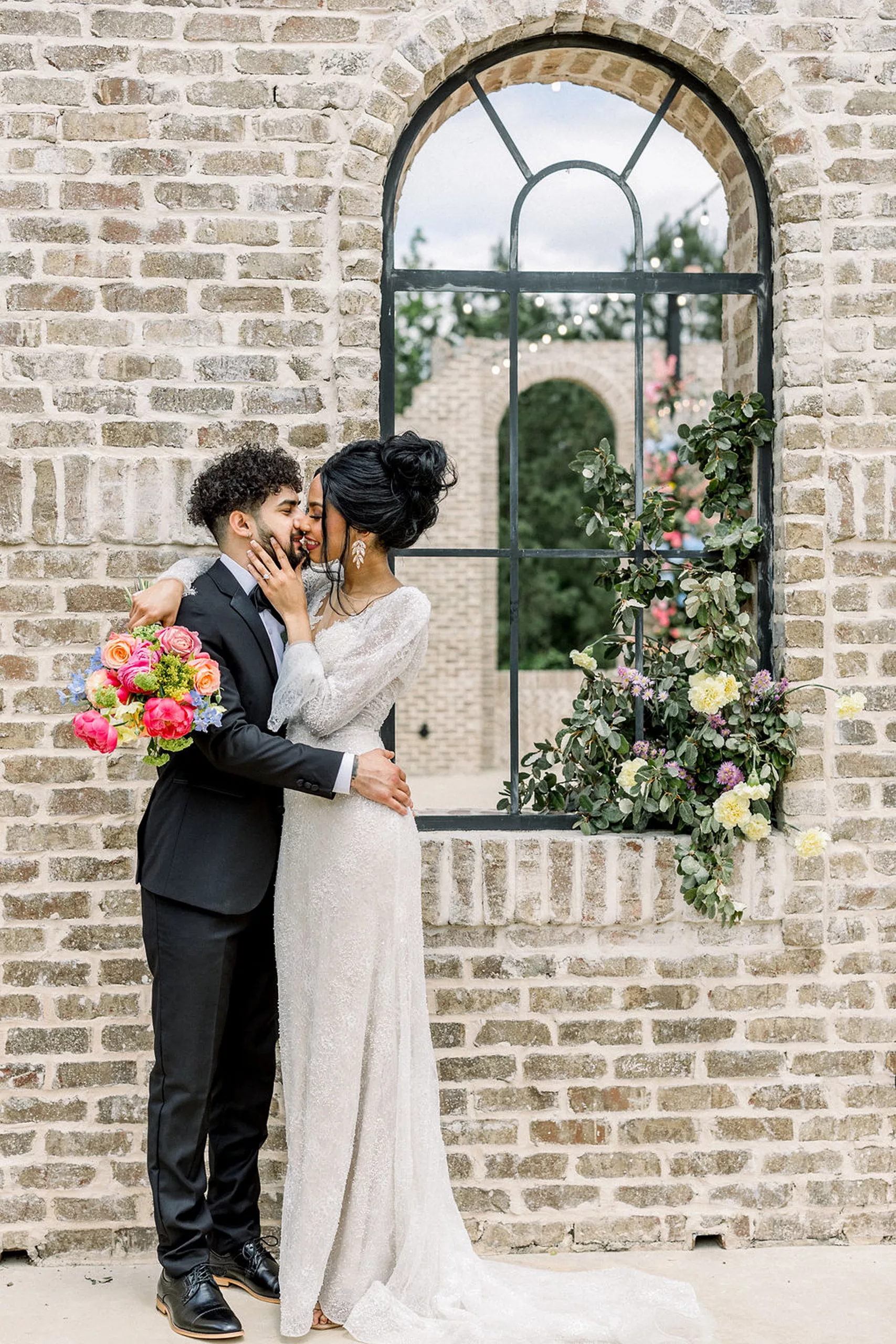 Newlyweds kiss while standing against a light brick wall with flowers in the window in a black suit and a white lace dress at the iron manor wedding venue