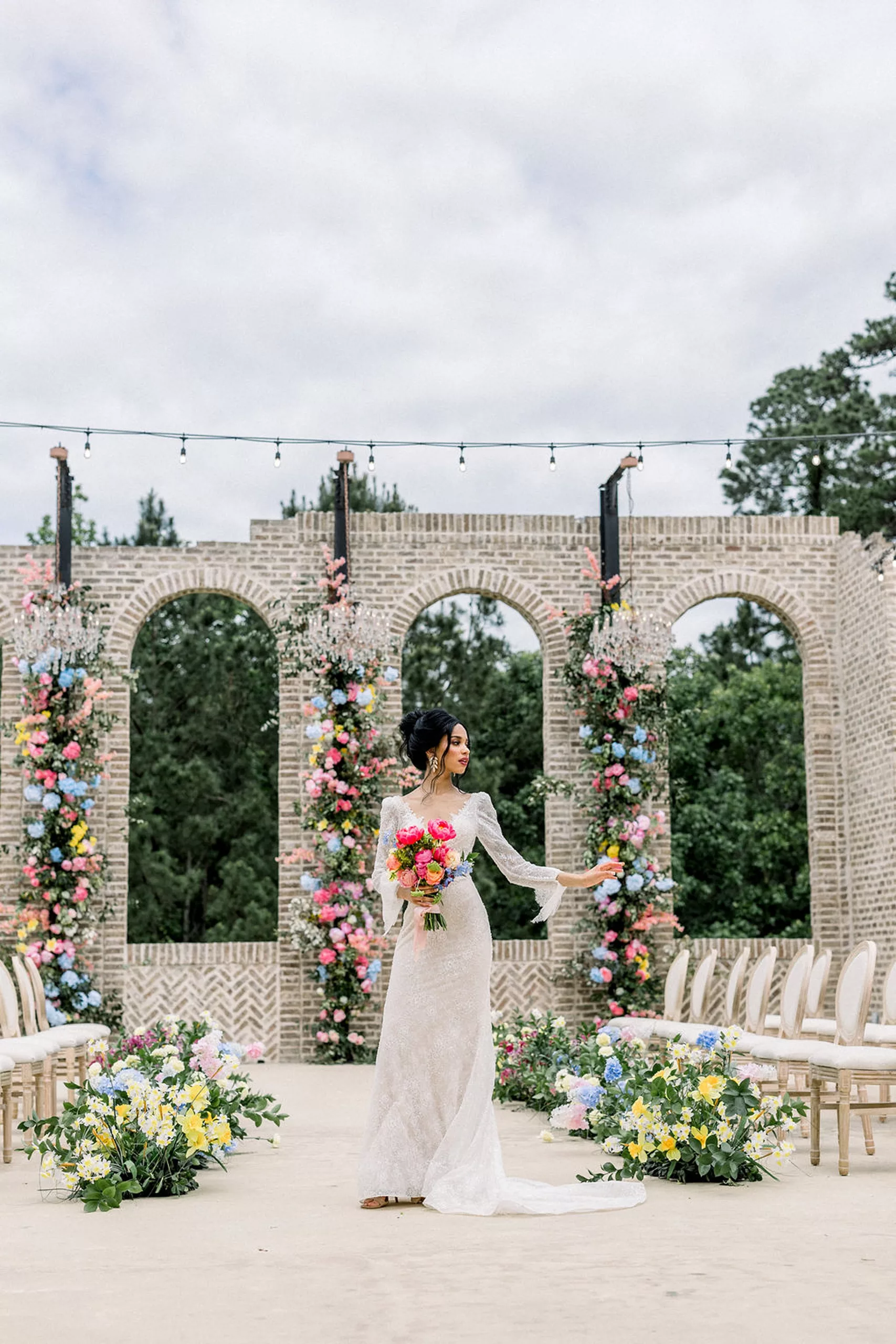 A bride in a white lace dress walks down the empty aisle of her outdoor iron manor wedding ceremony location with tall brick arches