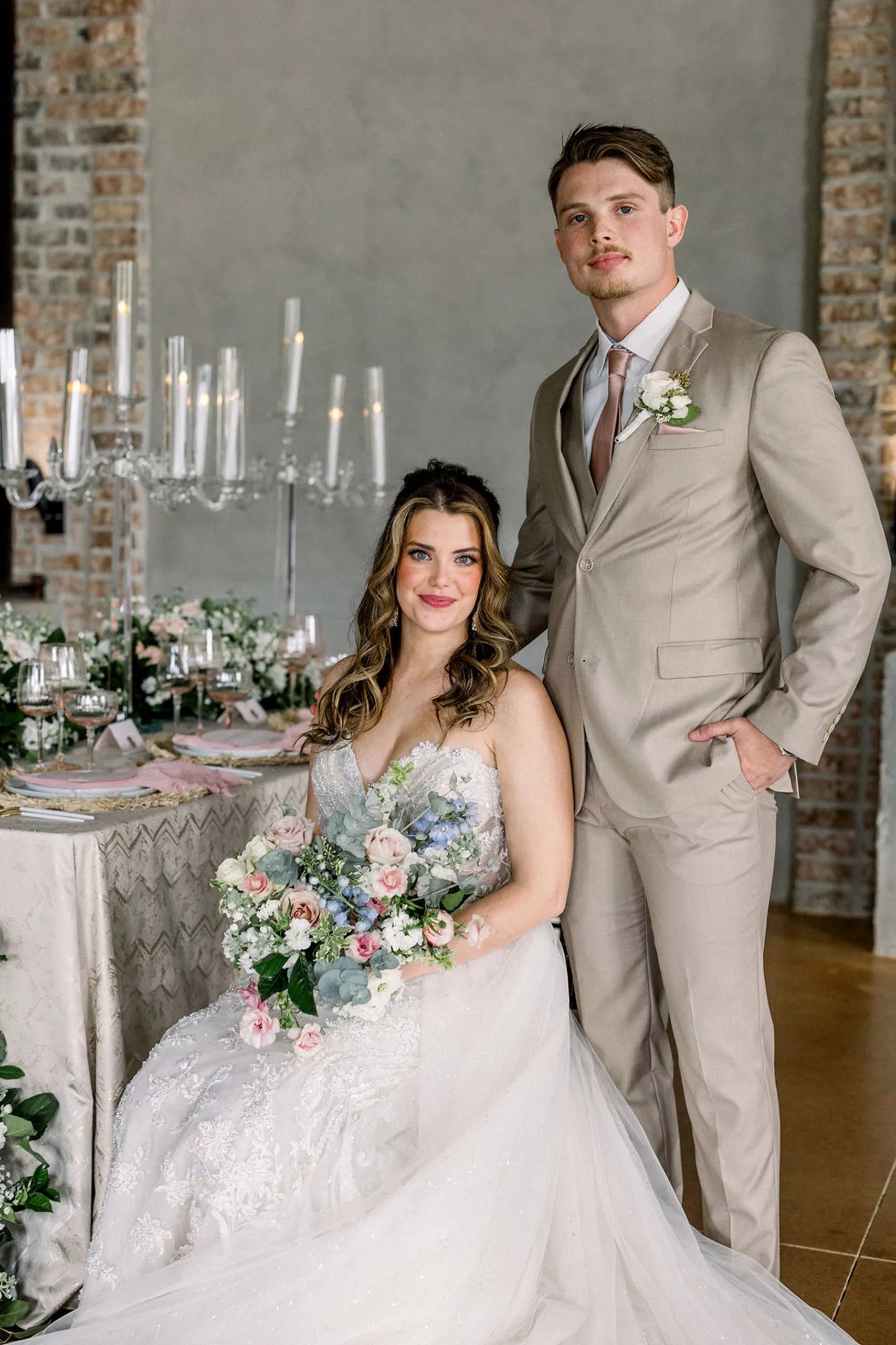 A bride in an embroidered dress sits in a chair at a reception table with her husband in a beige suit standing standing behind her with a hand on her shoulder at an iron manor wedding