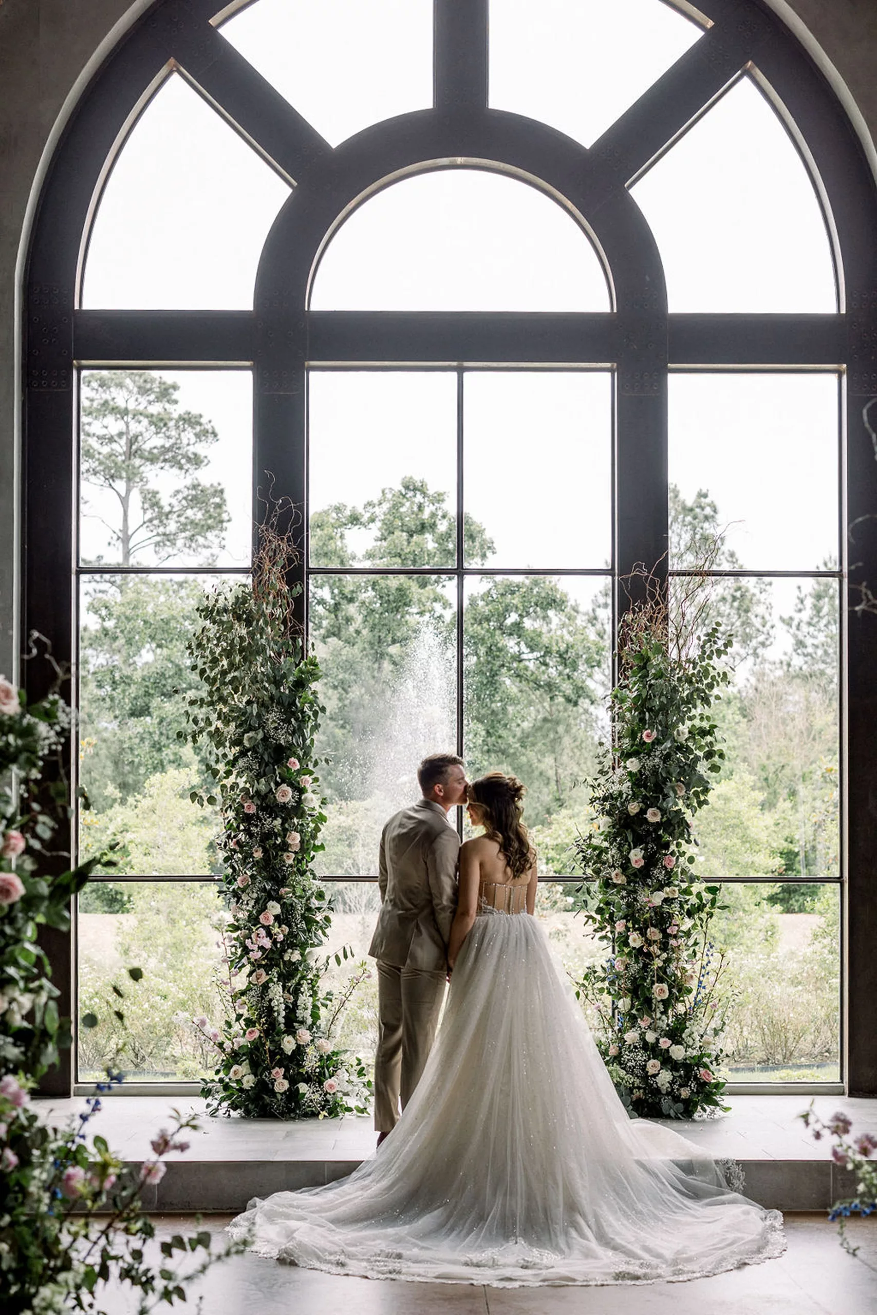 Newlyweds stand in a large window as the groom in a beige suit kisses his bride on the forehead at the iron manor wedding venue