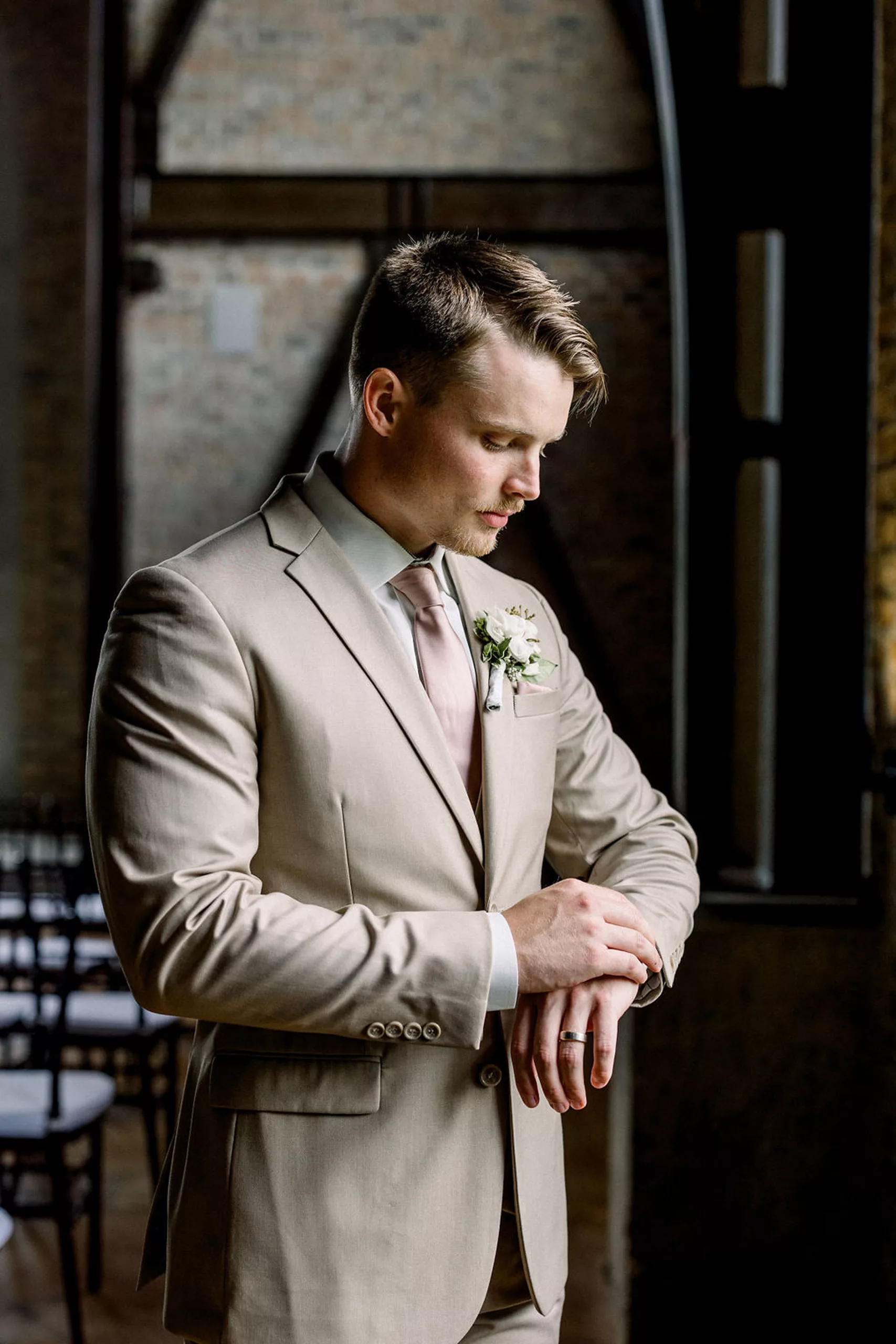 A groom checks his watch in a beige suit and pink tie in a window before his iron manor wedding