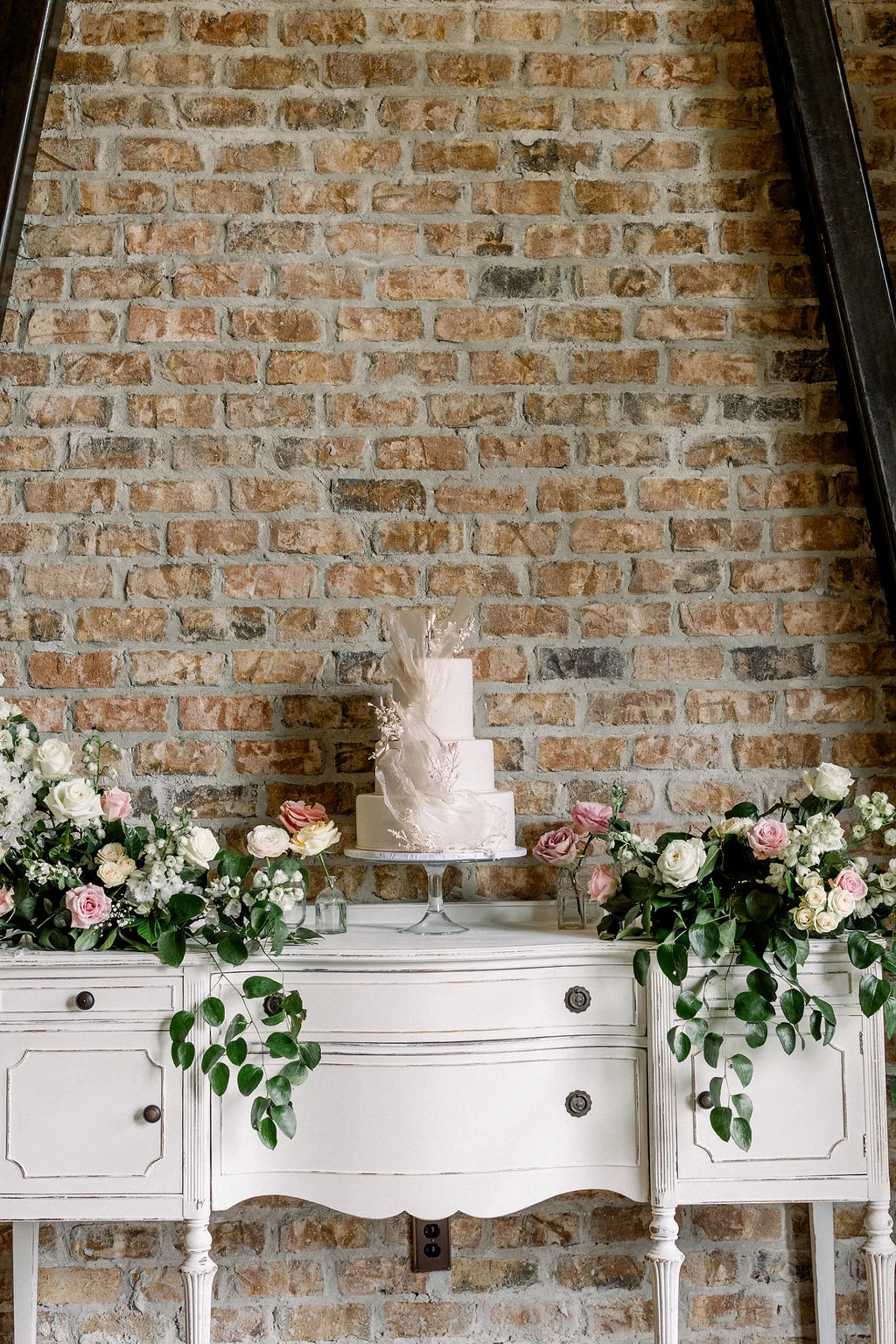 A three tier wedding cake sits on a table surrounded by pink and white roses at the iron manor wedding venue