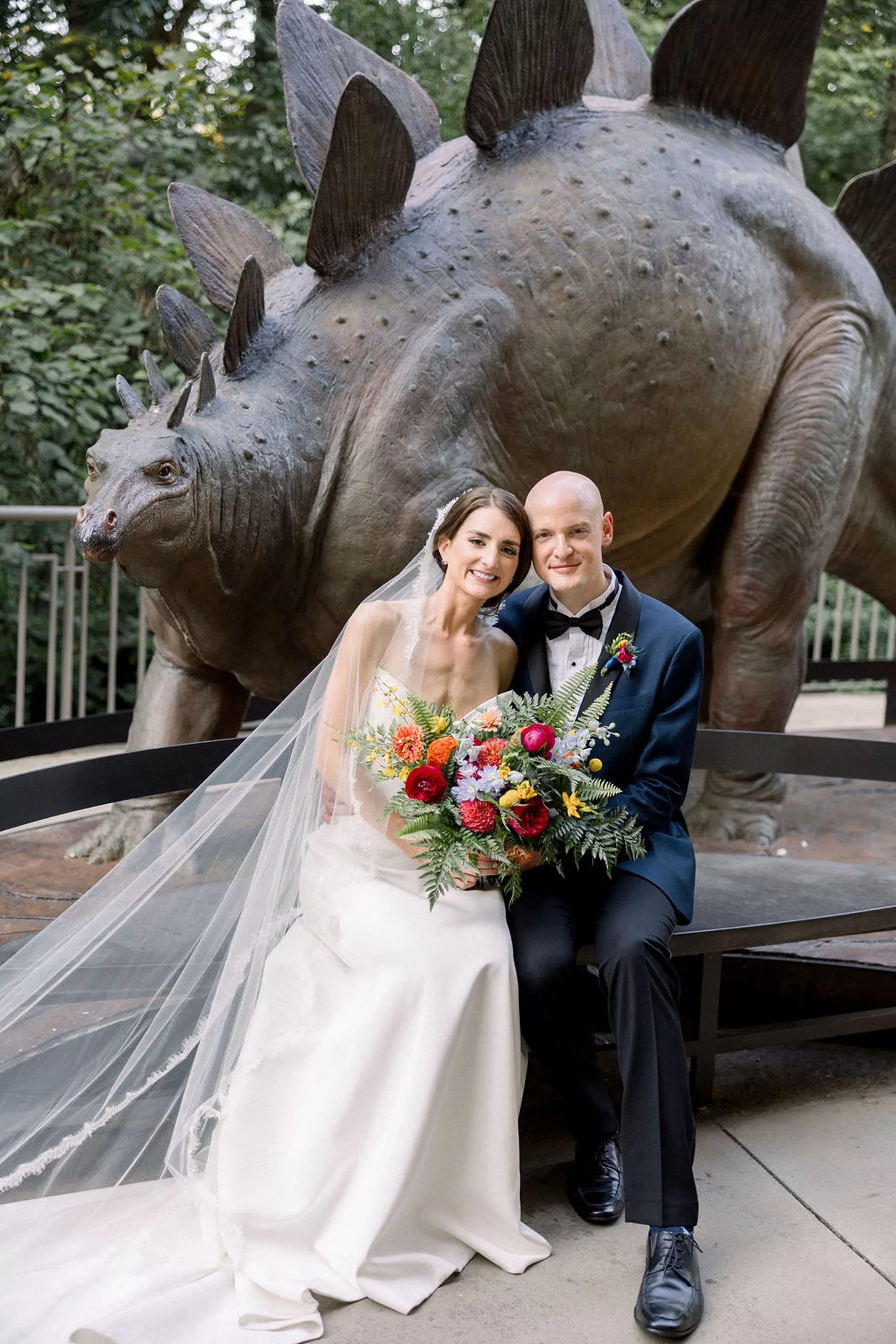 Newlyweds sit on a bench in front of a stegosaurus statue at their jurassic park wedding