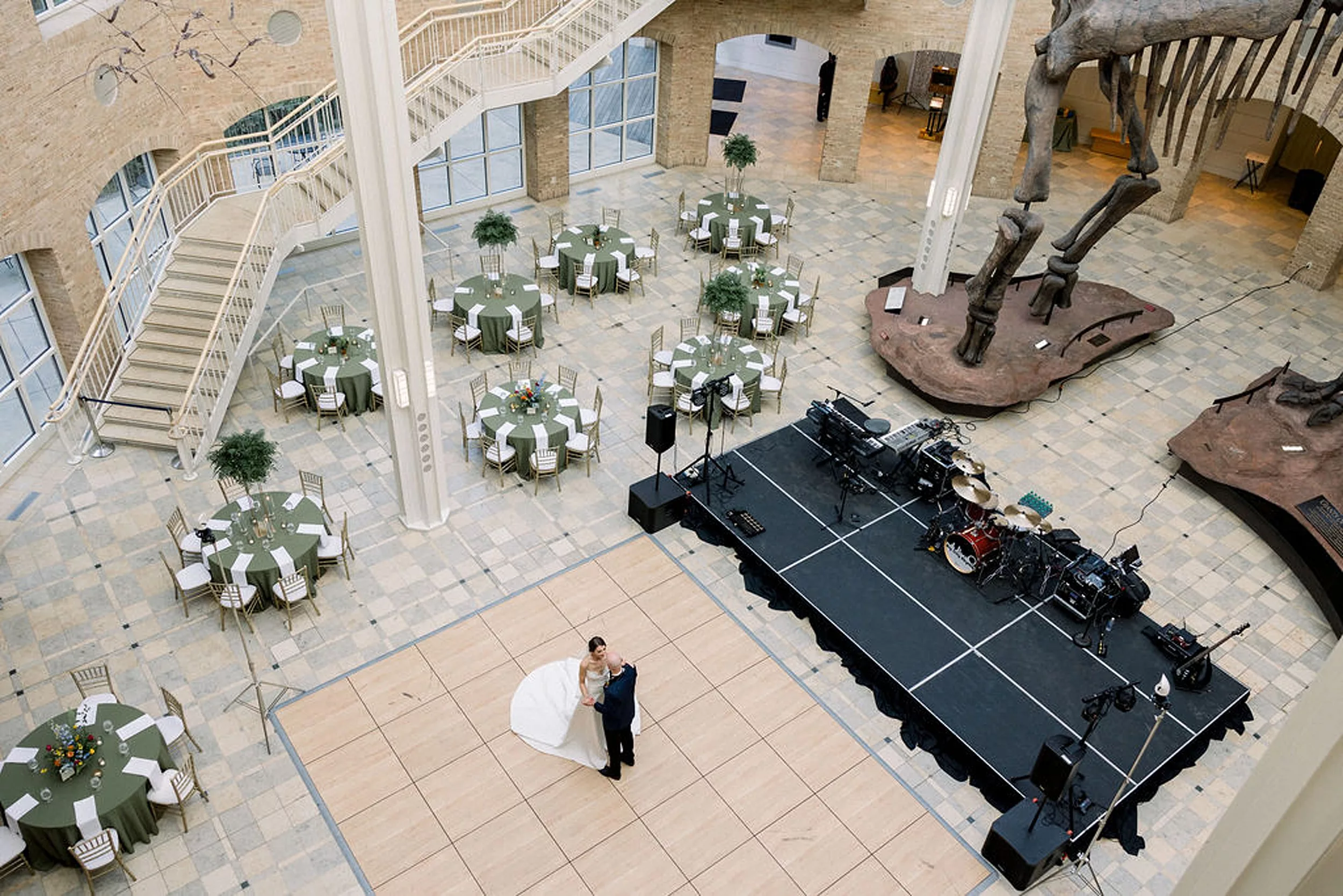 A bride and groom dance alone in an empty reception room under a giant dinosaur skeleton
